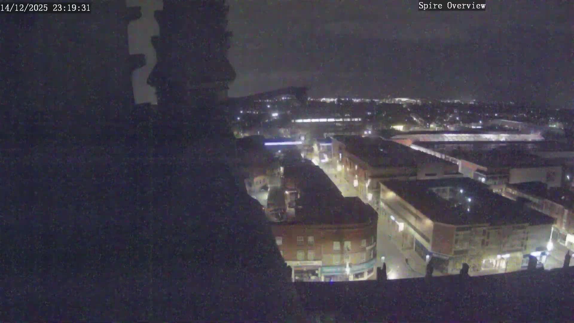 The image captures an overcast day overlooking an urban landscape from a moss-covered stone architectural structure, revealing a dense mix of traditional brick buildings, modern commercial complexes, and a large shopping center under a cloudy sky.