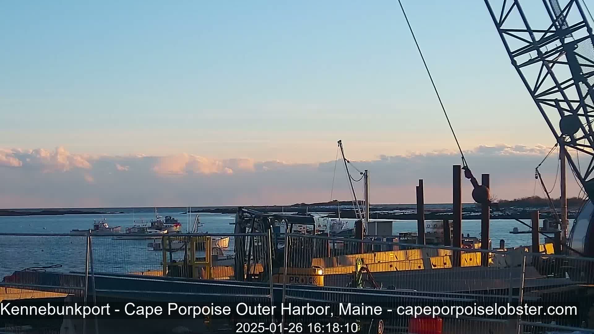 Under a partly cloudy sunset sky, a harbor scene shows several boats, construction equipment, and a large crane at a waterfront construction site.