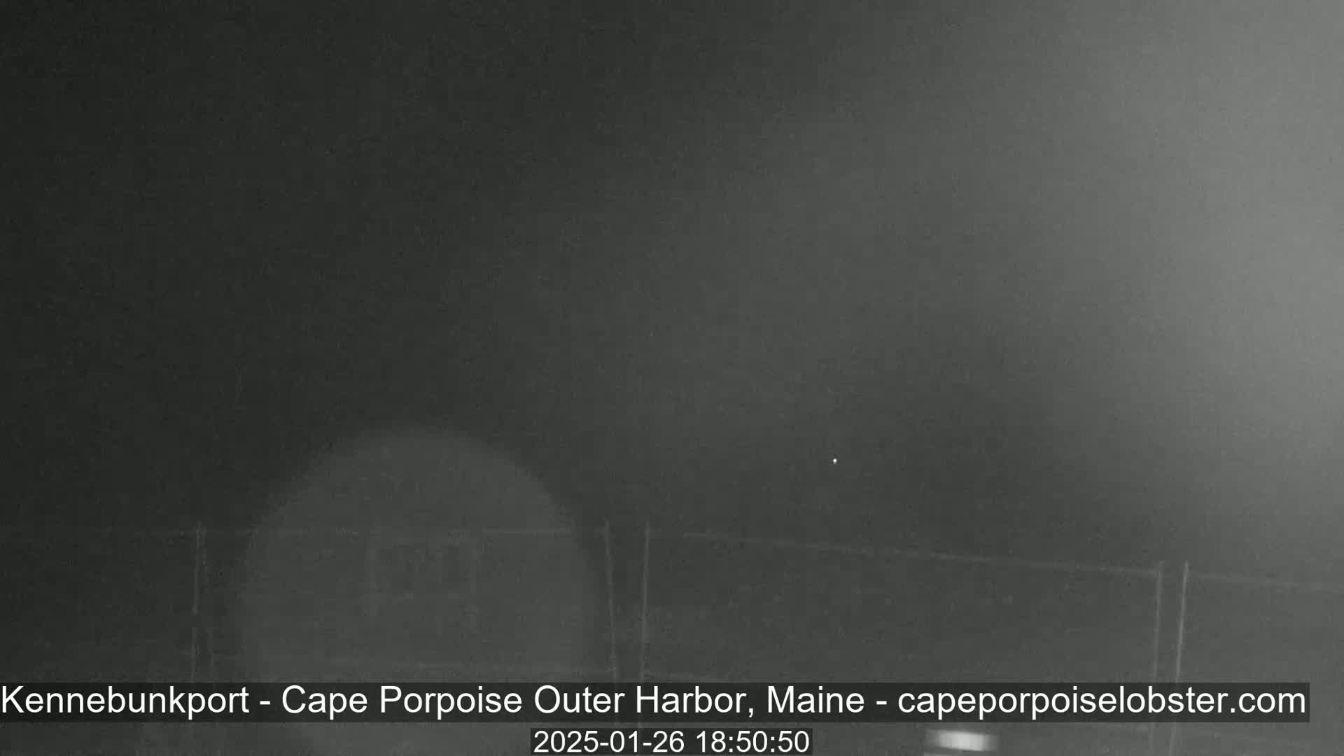 A nighttime, blurry outdoor image shows a dark, possibly foggy or hazy scene with a fence in the foreground.