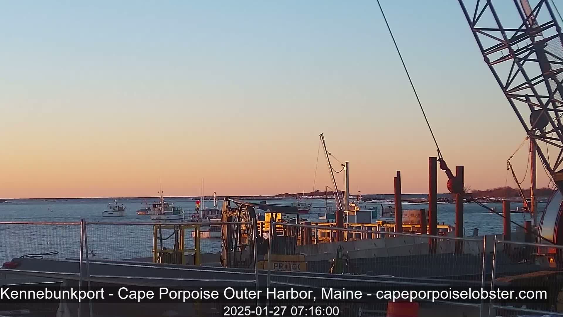 At sunset, several boats are moored at a harbor, with a large crane and other harbor equipment visible in the foreground.