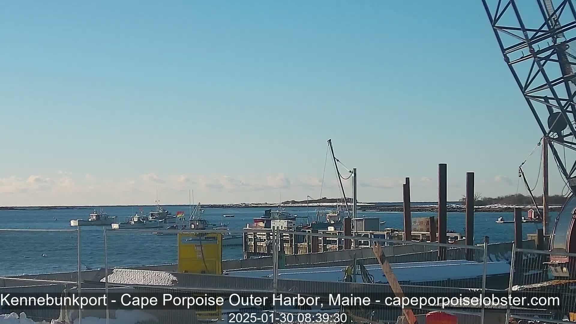 Several fishing boats are moored in a harbor on a partly cloudy day with some snow visible on the ground.