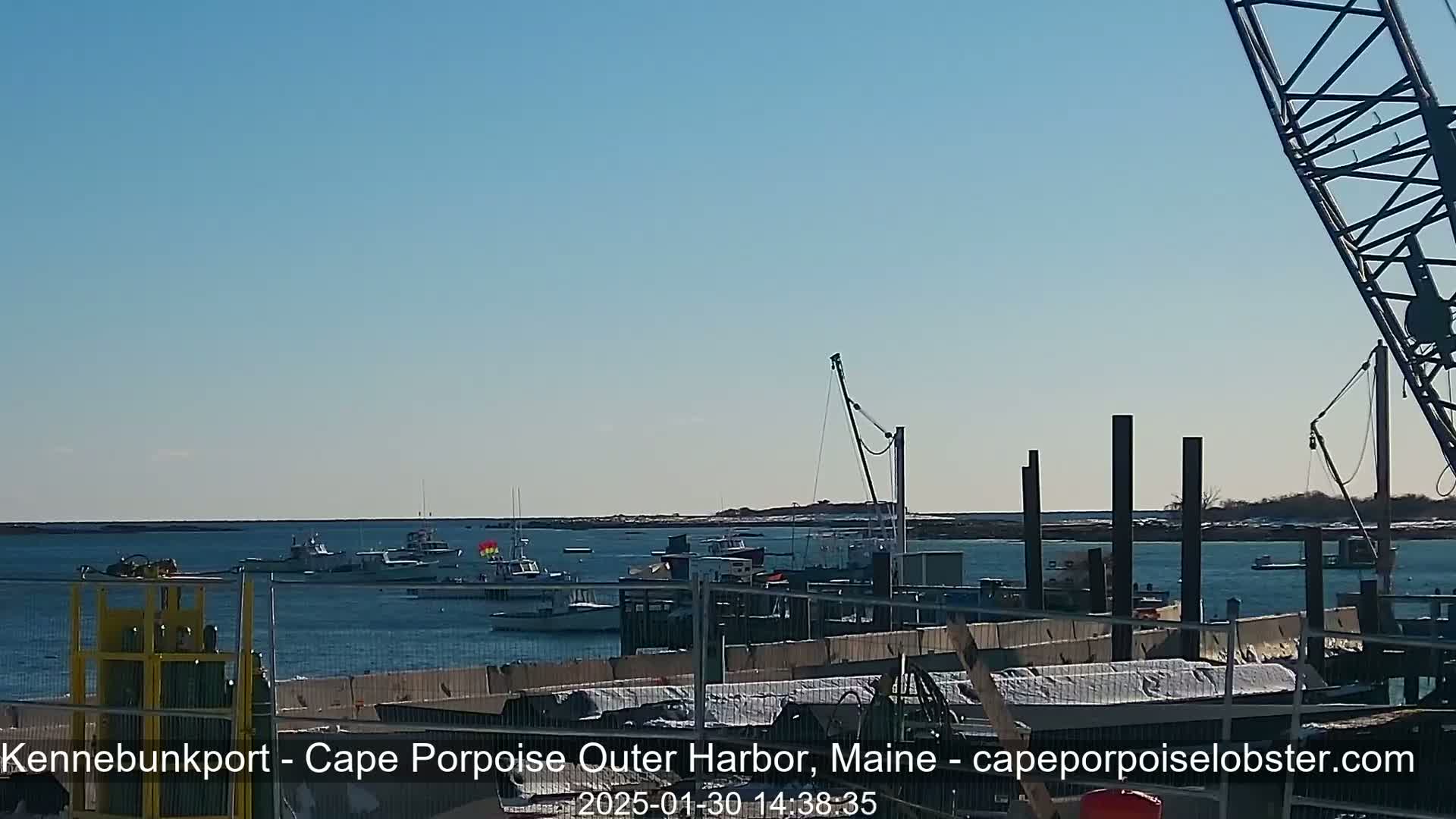 Several fishing boats are moored in a harbor under a clear blue sky, with a crane and construction elements visible on the dock.