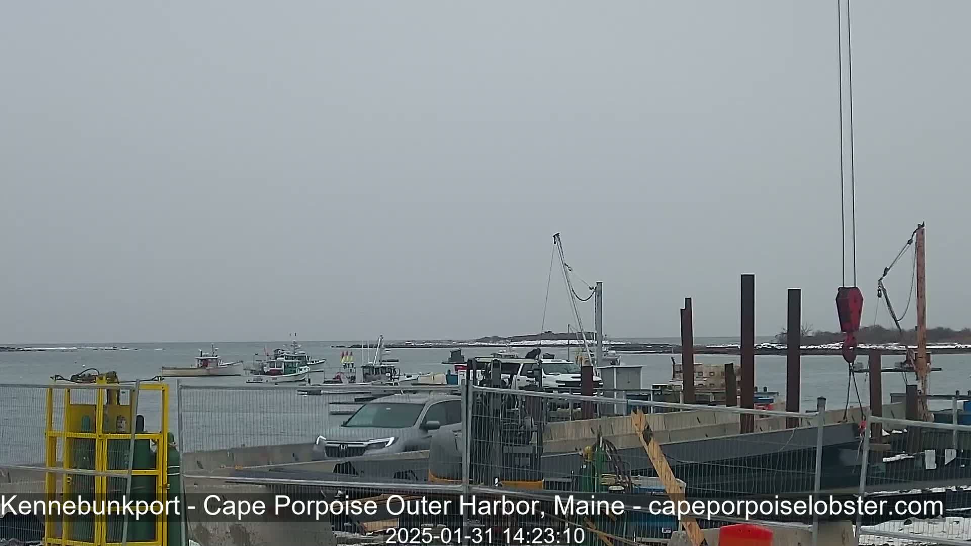 Several fishing boats are moored in a harbor under an overcast sky, with construction work and vehicles visible on the docks.