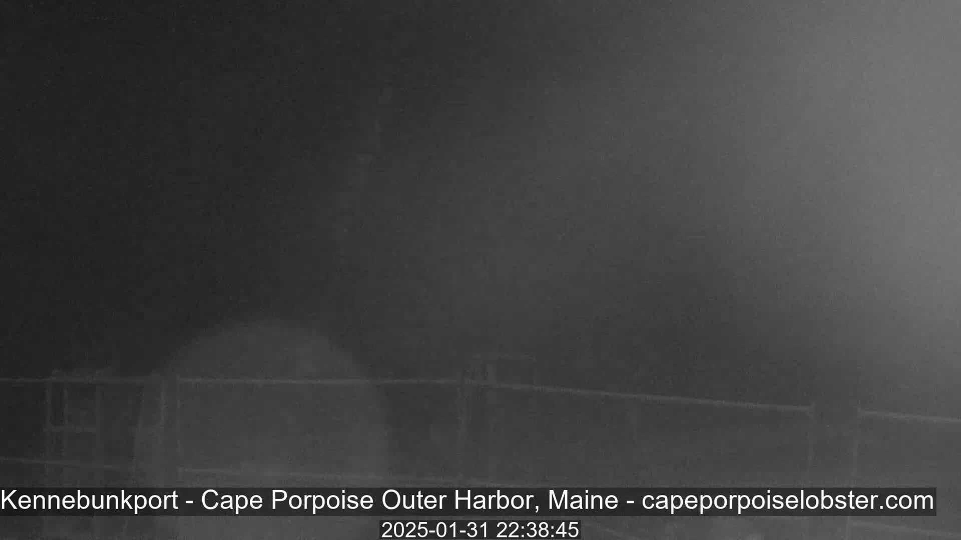 A dark, blurry nighttime image shows a fence and possibly a building in foggy or misty conditions.