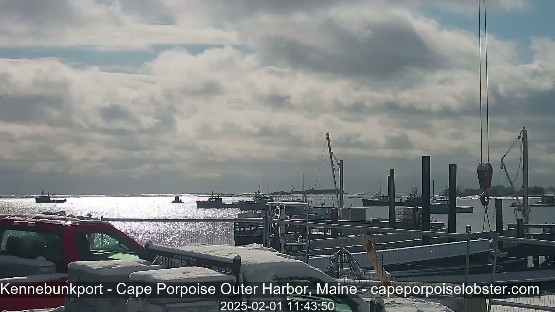 A partly cloudy day shows several boats in a harbor, with snow on the docks and a red truck parked nearby.