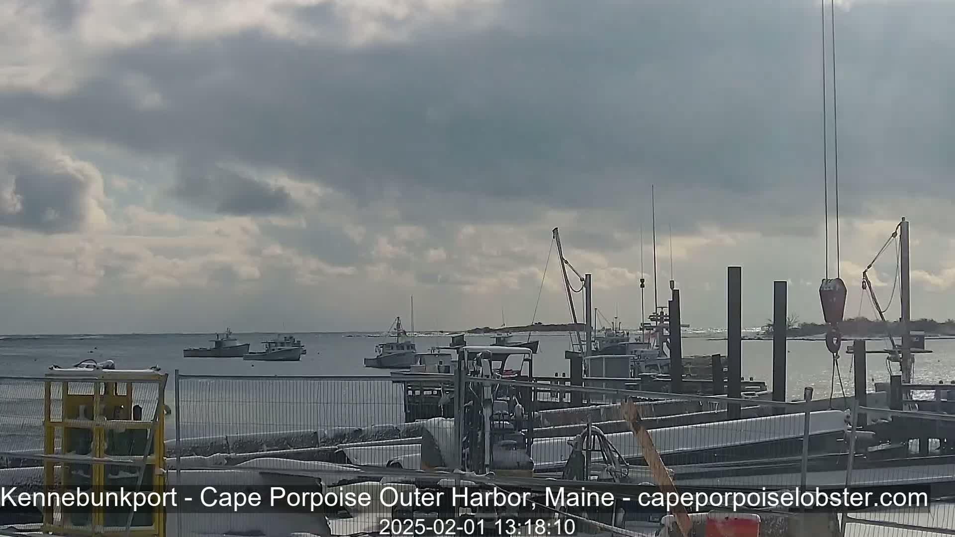 Several fishing boats are moored in a harbor under a cloudy sky, with snow on the ground in the foreground.