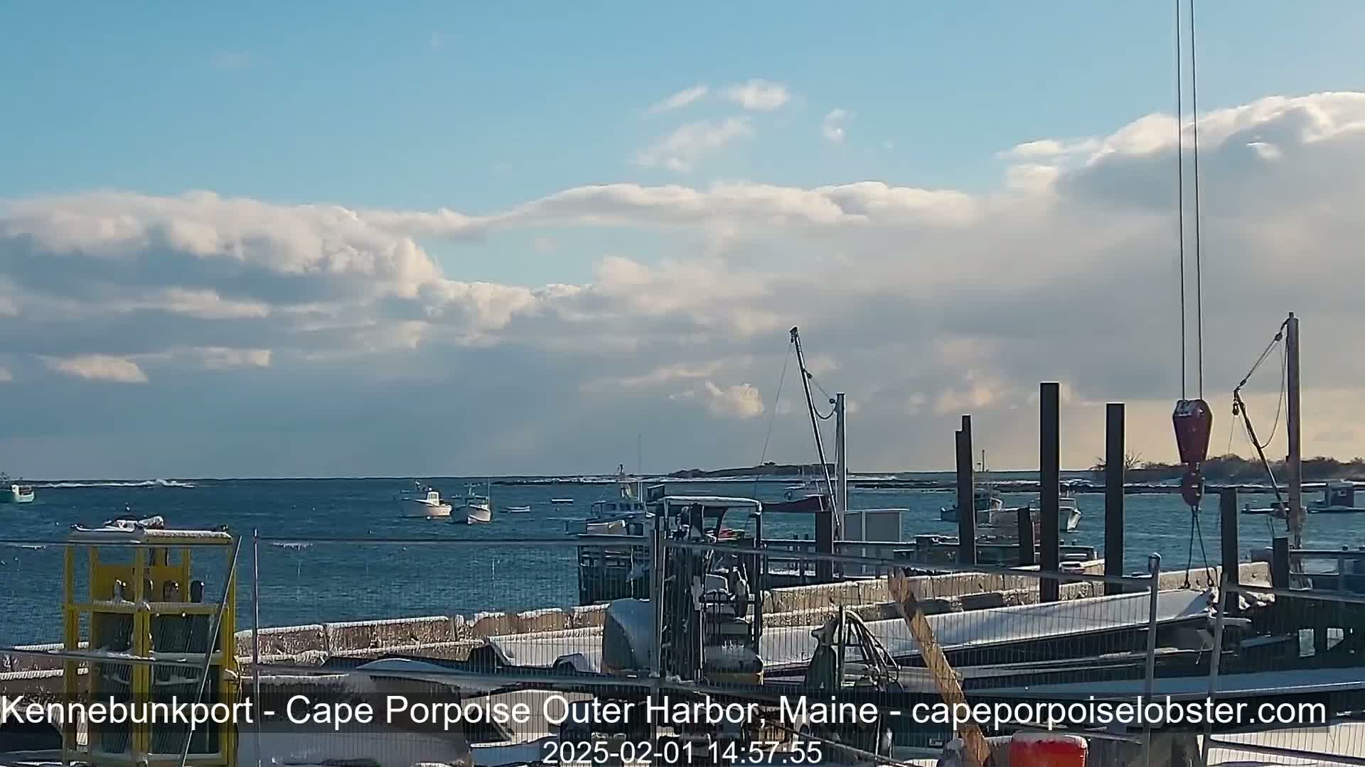 A partly cloudy sky overlooks a harbor filled with several boats, some docked and others in the water, with snow visible on the ground and some docked boats.