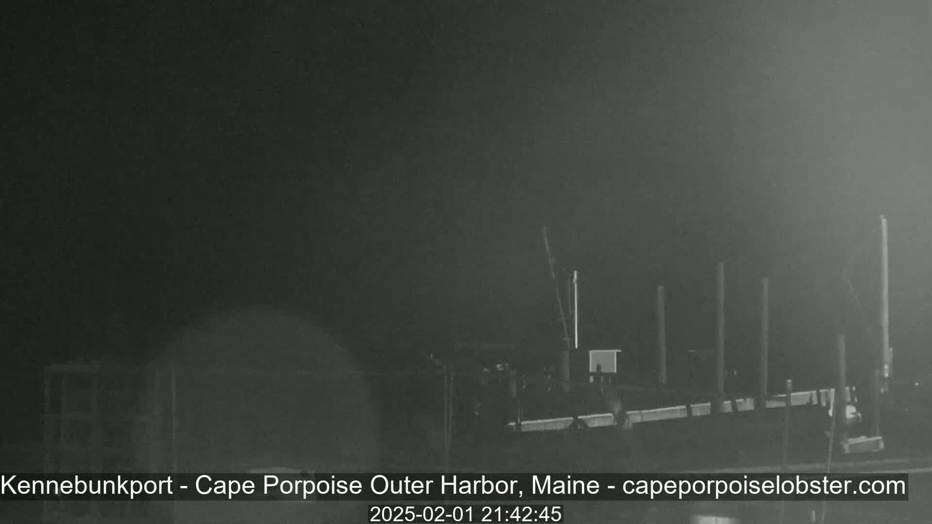 A nighttime, low-visibility view of a harbor, showing several dark structures and poles extending from the water.
