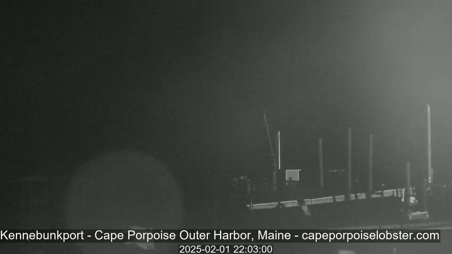 A nighttime, low-light image shows a dark harbor with several tall, thin structures and a large, dark mass that appears to be a boat or dock.