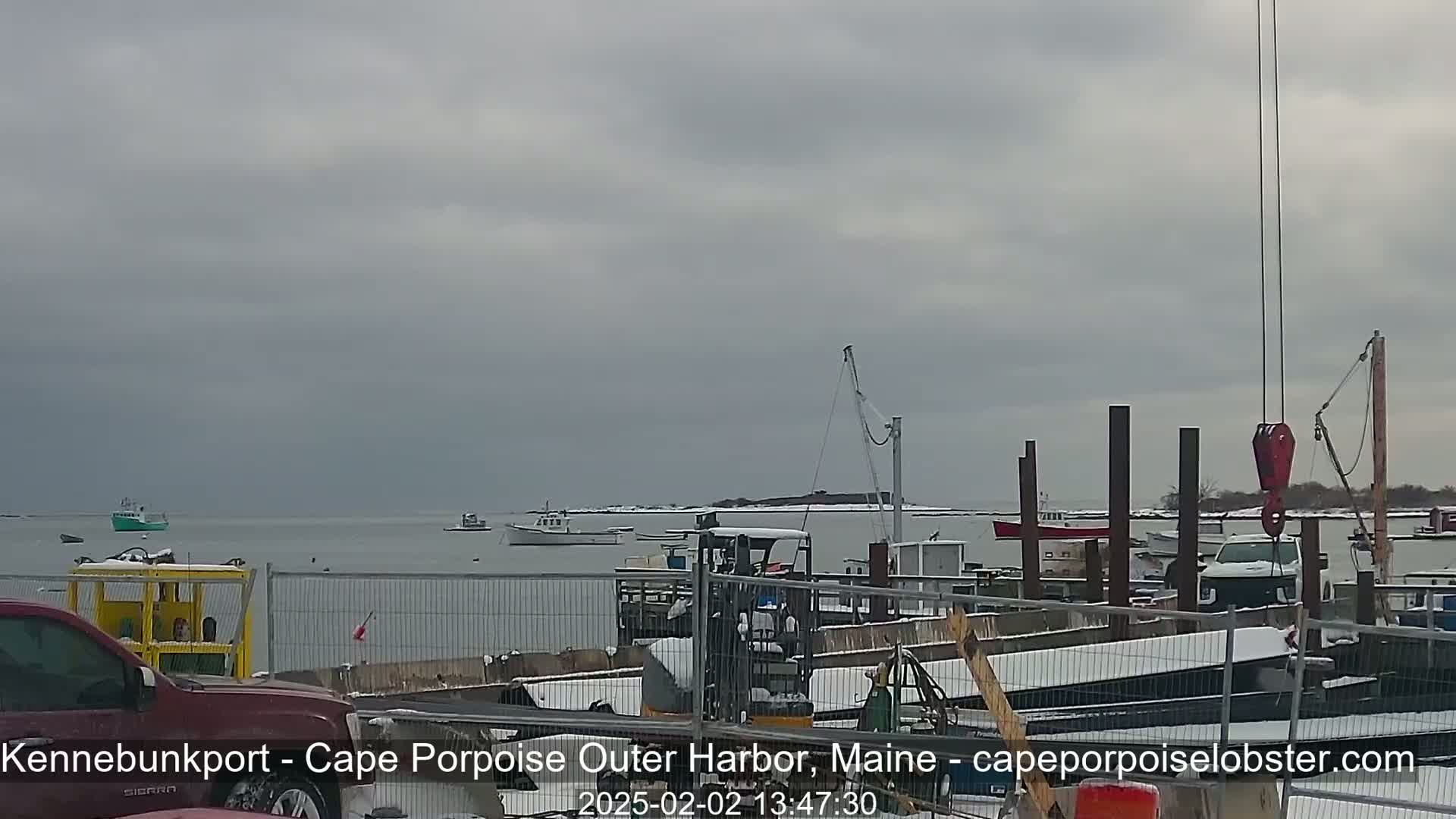 A harbor scene on an overcast day shows several boats on the water and a dock with construction materials and equipment, including a forklift and a crane, covered in a light layer of snow.