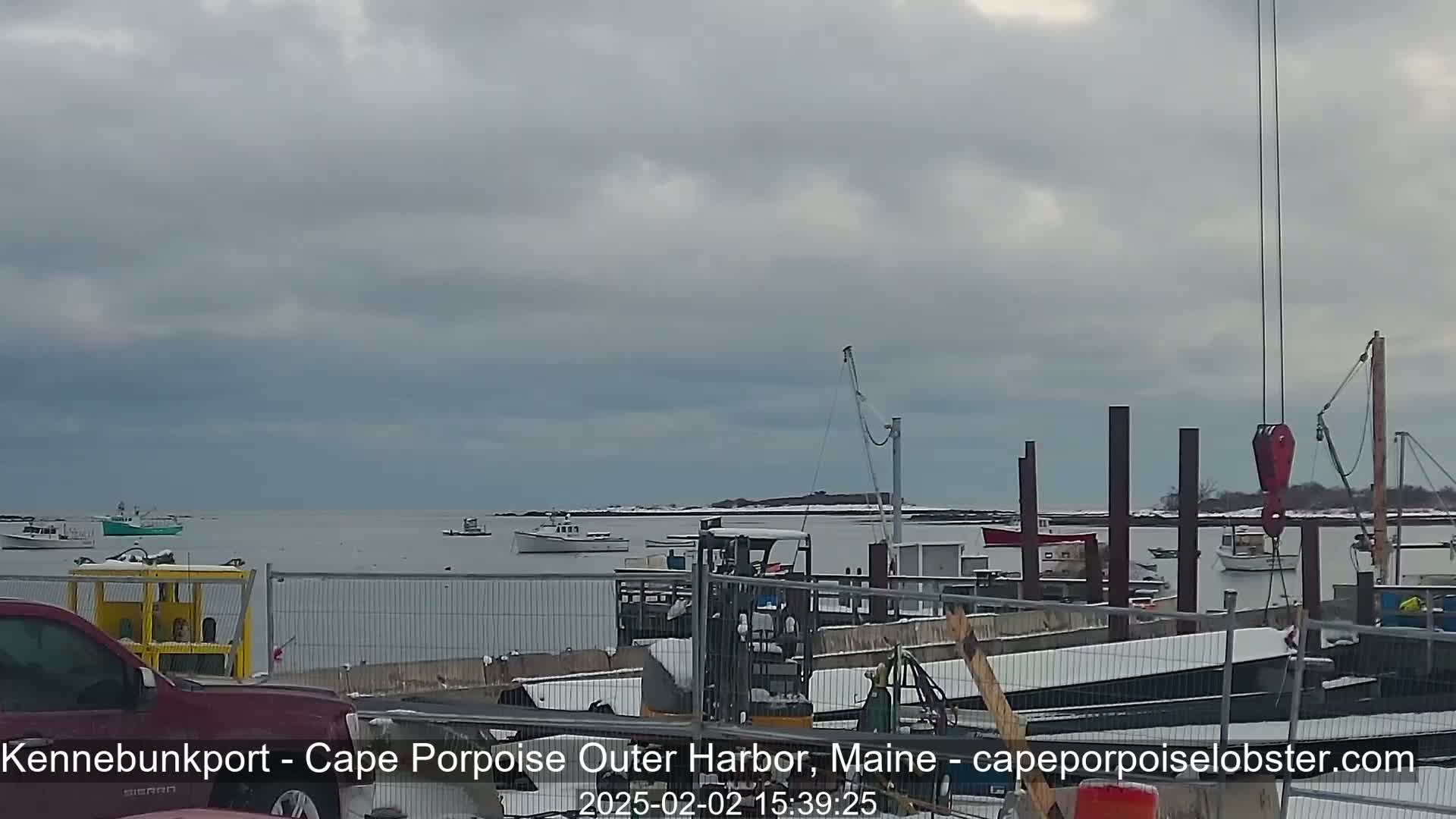 A harbor scene shows several boats in calm water under an overcast sky, with snow on the ground and some construction or maintenance work underway at a dock.