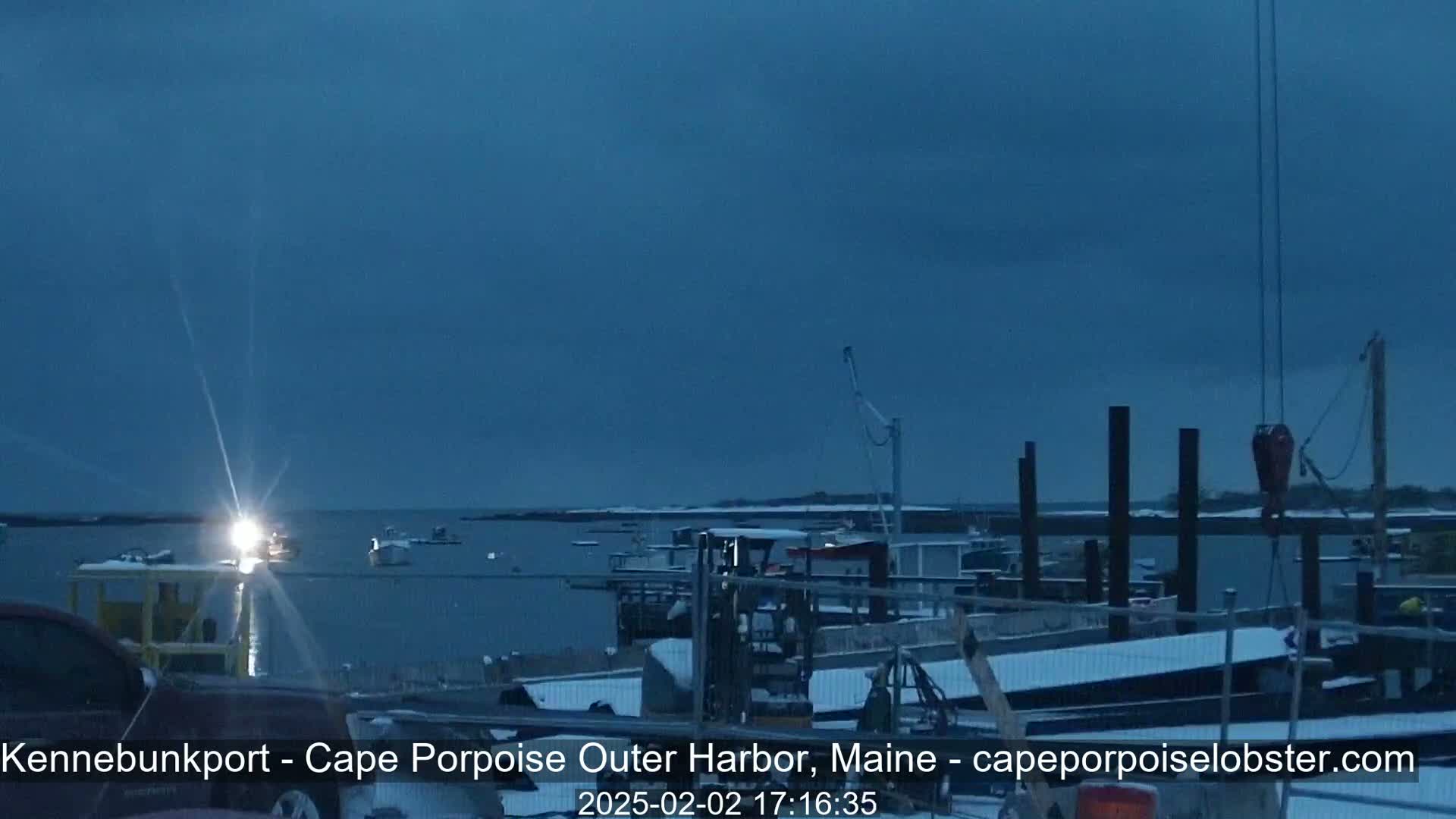 A snow-dusted harbor at twilight shows boats moored at docks under a dark, overcast sky.