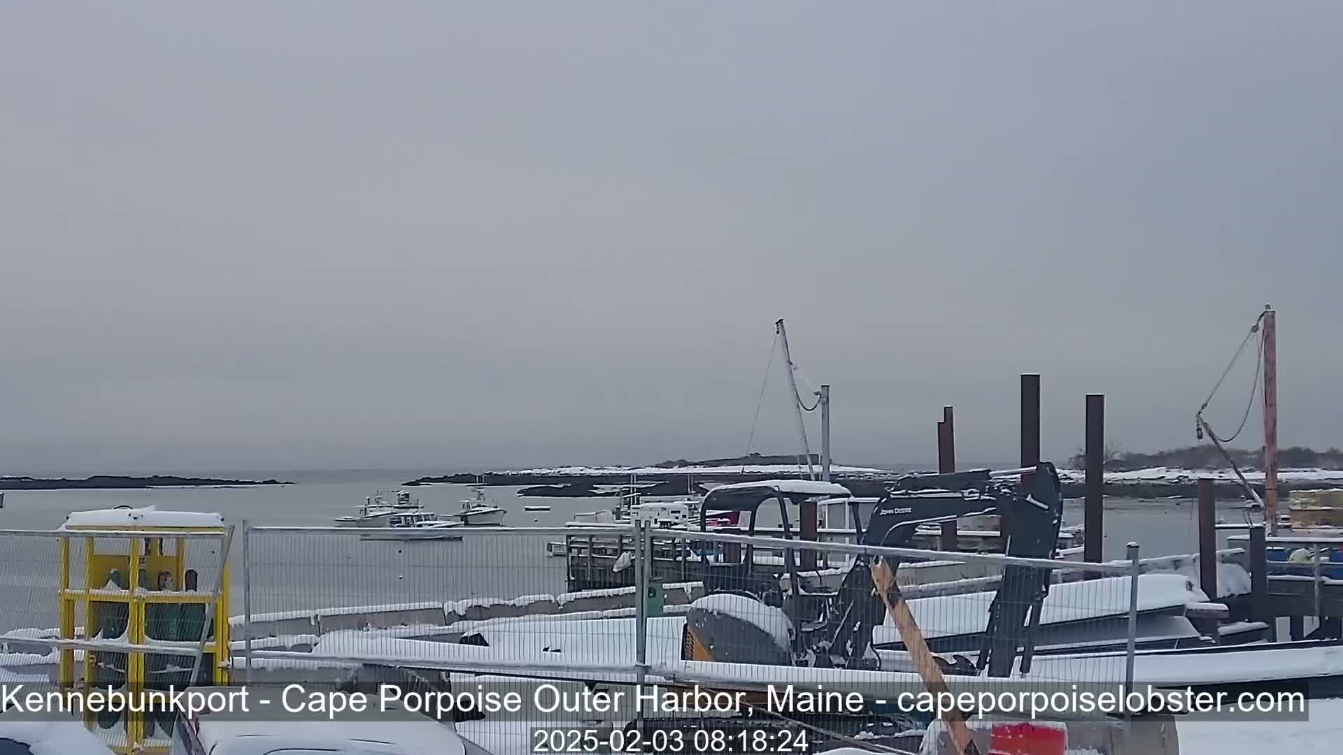 A snow-covered harbor scene shows several boats, a small excavator, and other equipment behind a chain-link fence.