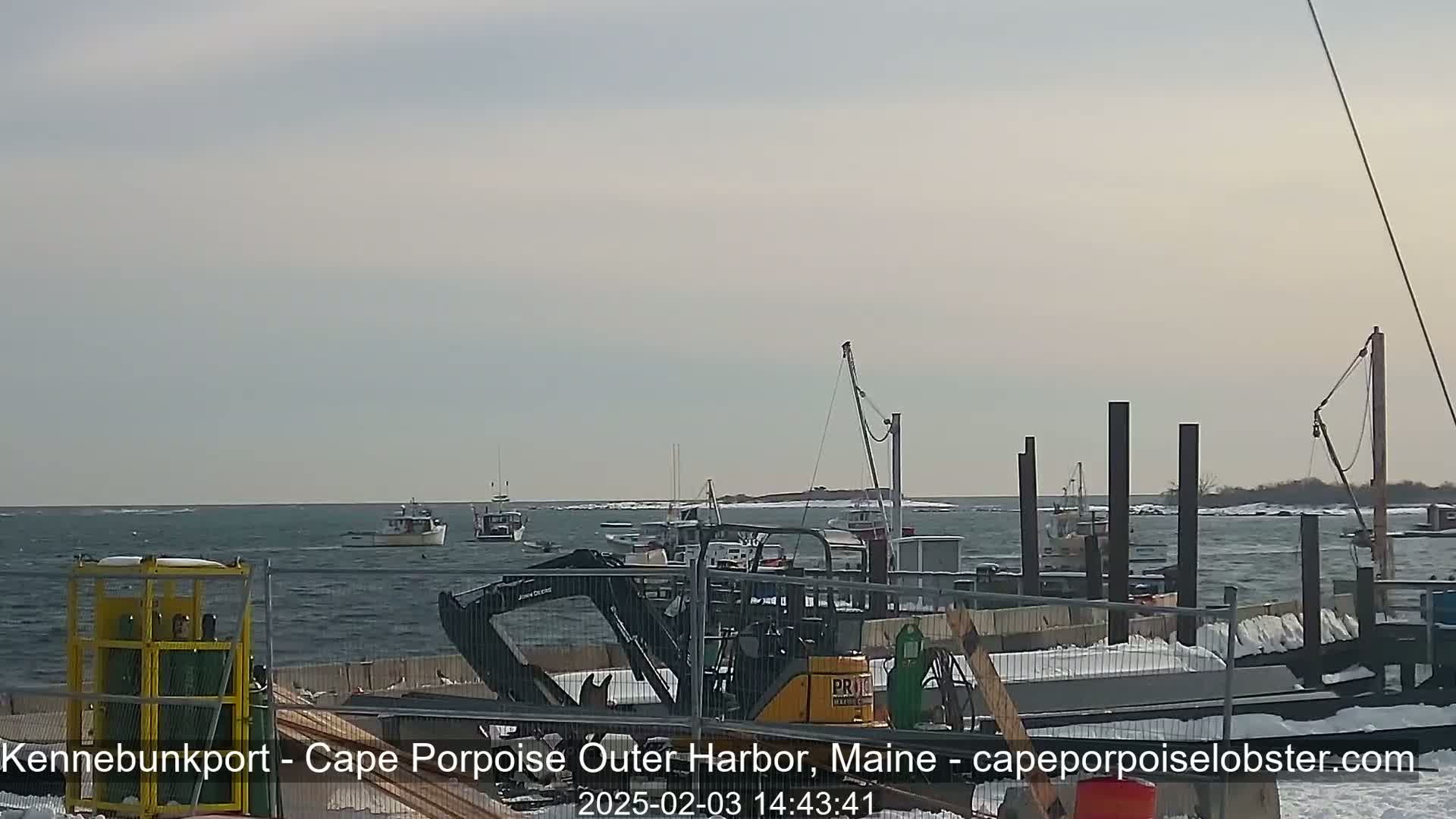 Several fishing boats are in a harbor with a partly cloudy sky and a light chop on the water, and construction equipment is visible on a nearby dock with snow on the ground.