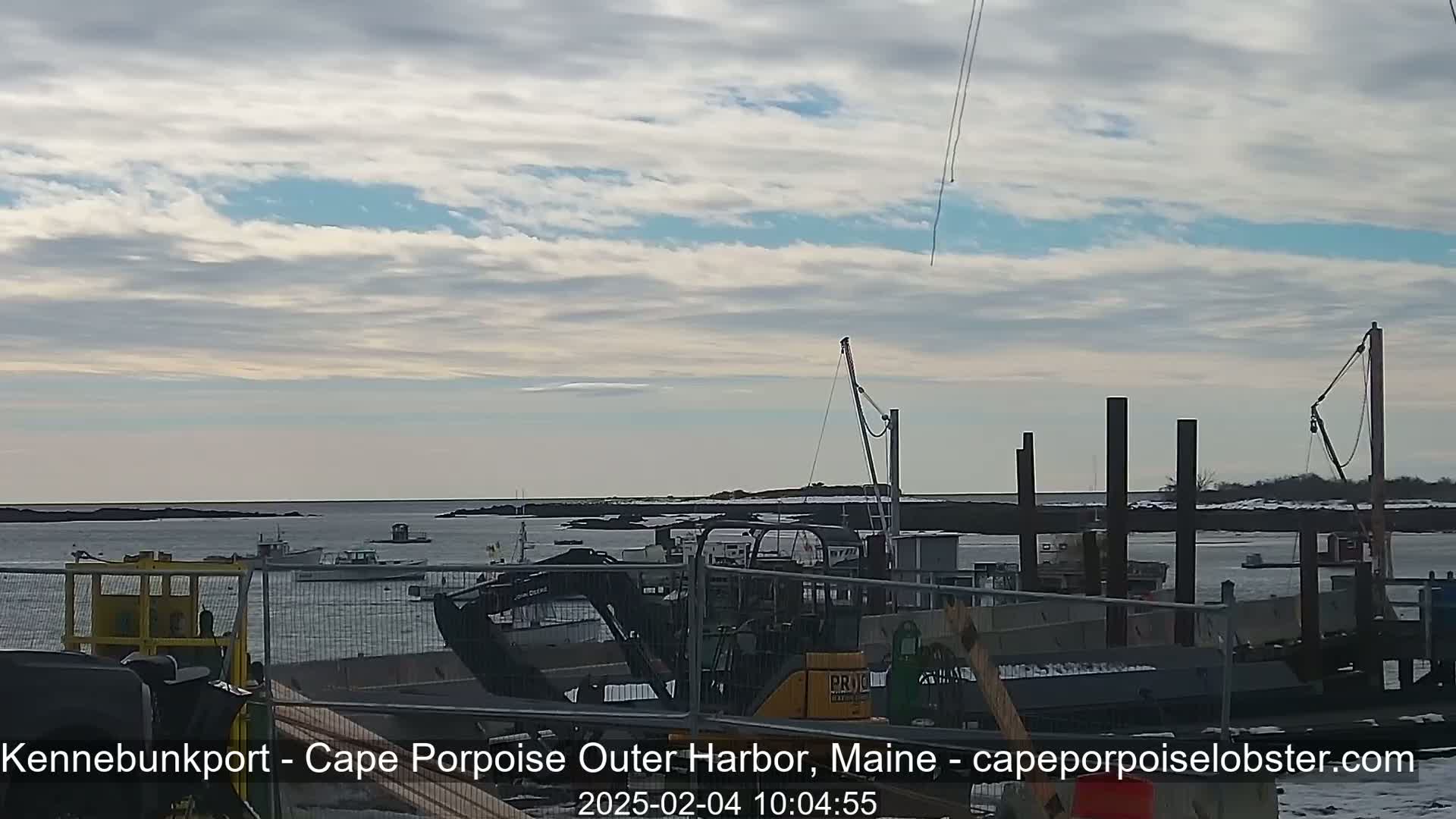 A harbor scene under a mostly cloudy sky shows several boats moored near a construction site with heavy machinery.