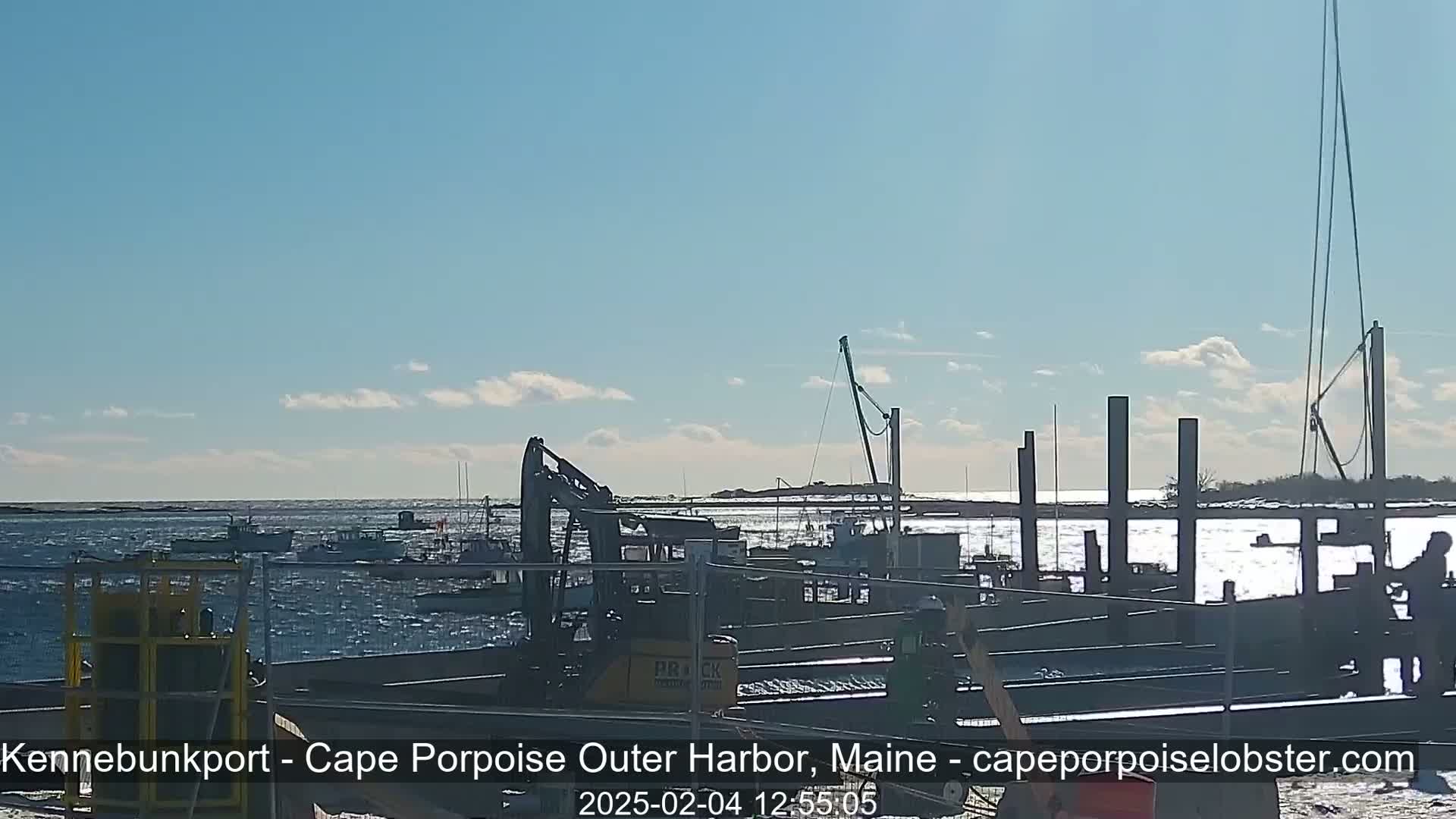Under a mostly sunny sky, numerous fishing boats are docked at a harbor, with construction or maintenance work underway near a crane and some workers present.