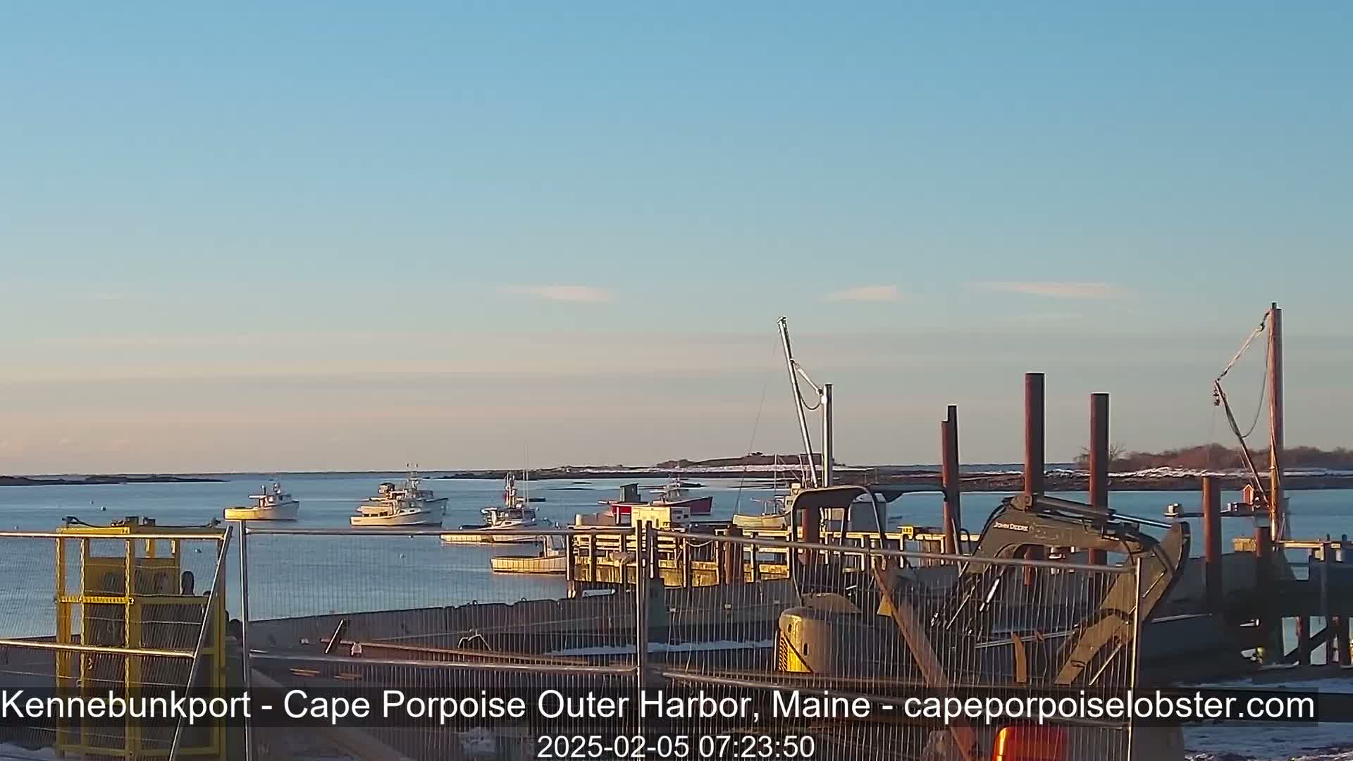 Several fishing boats are moored in a calm harbor under a clear, pale blue sky, with construction equipment visible on a nearby dock.