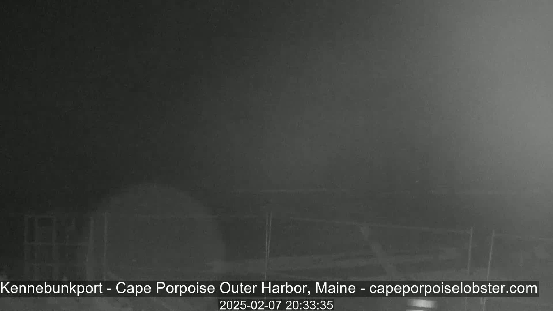 A low-light, nighttime image shows indistinct dark shapes, possibly structures or vehicles, in hazy or foggy conditions.