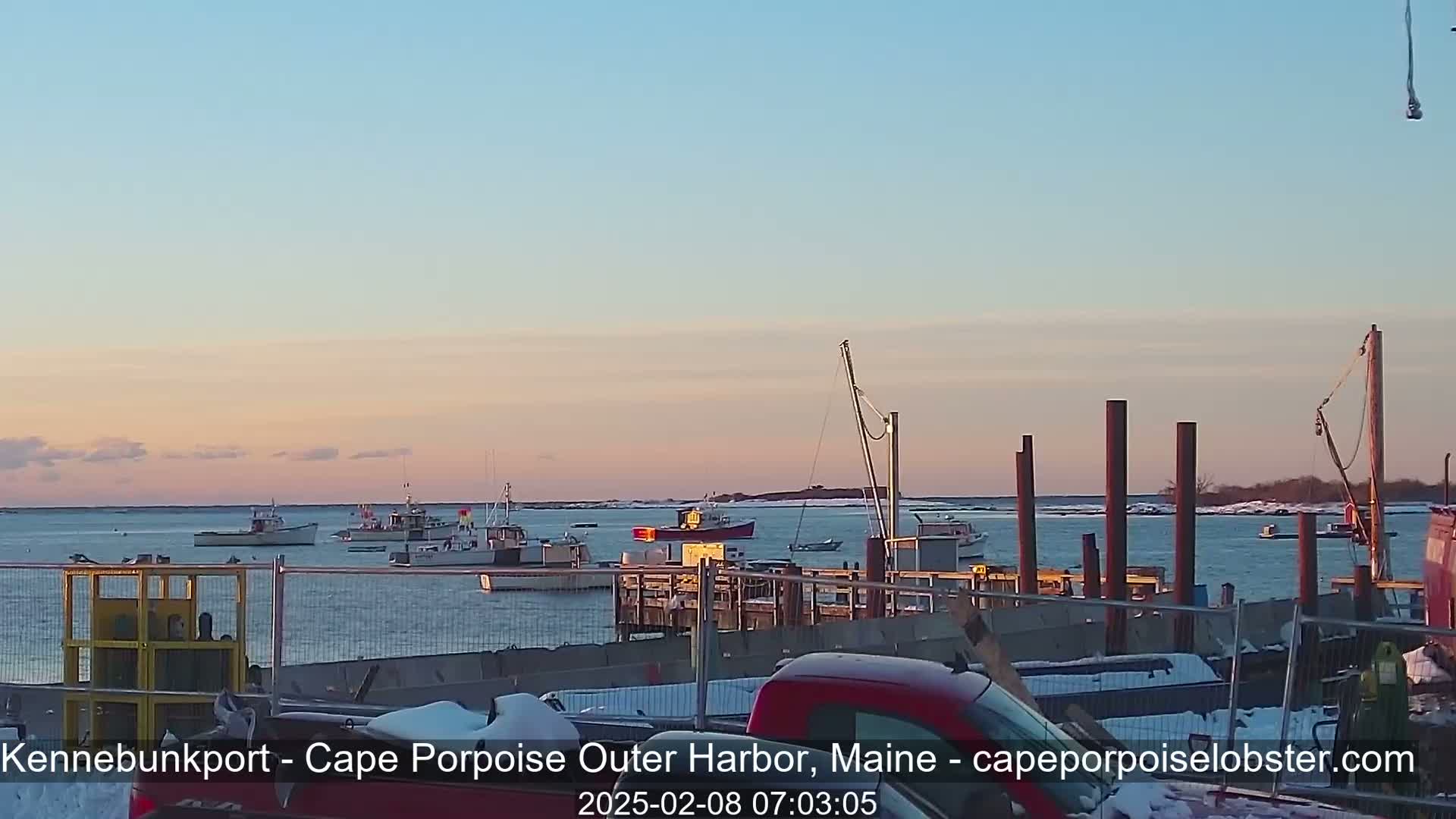 Several fishing boats are moored in a harbor at sunset under a clear, pale pink sky, with a small amount of snow visible on the ground near the docks.