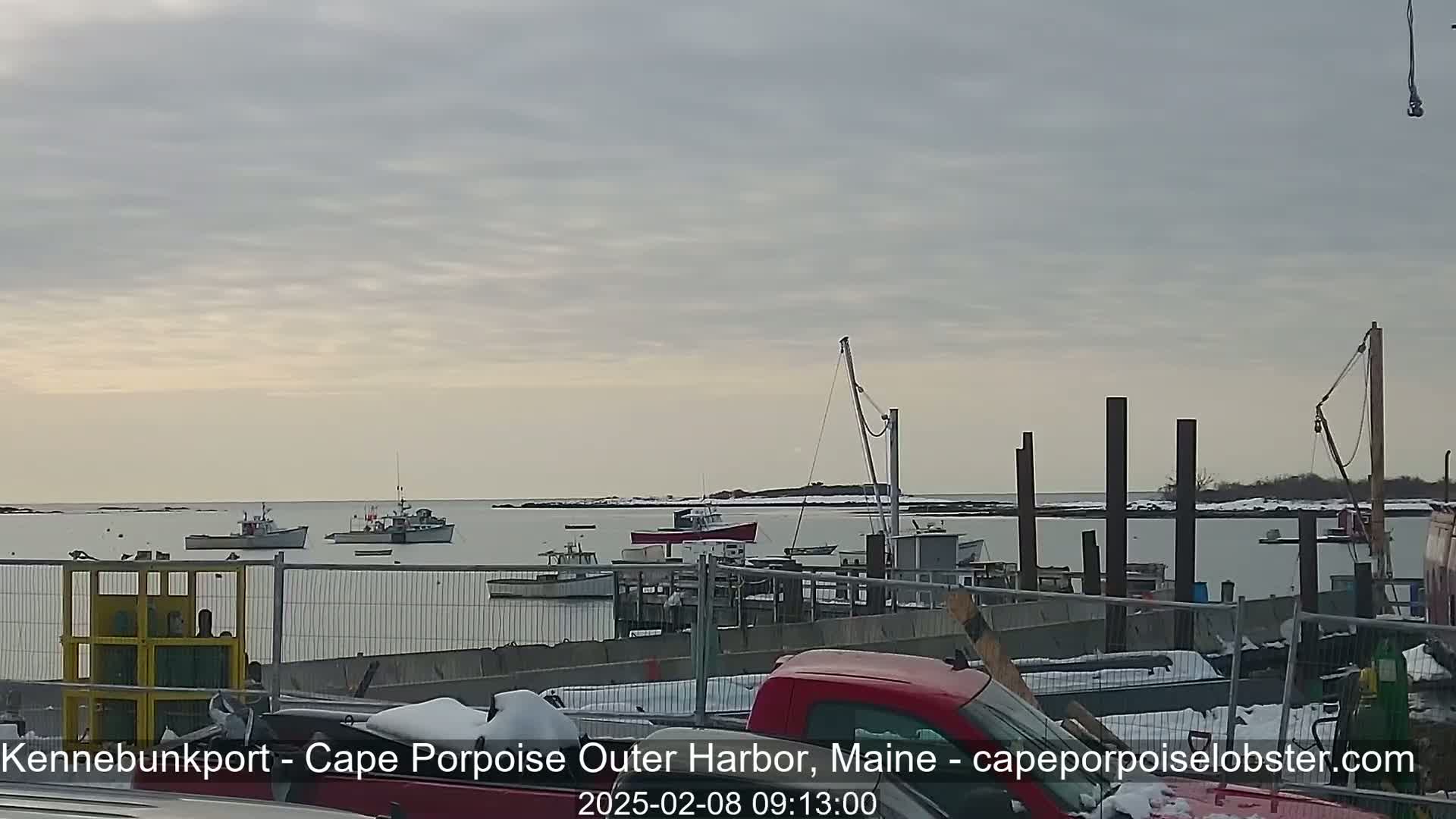 Several fishing boats are moored in a harbor under an overcast sky, with some snow visible on the ground and parked vehicles.