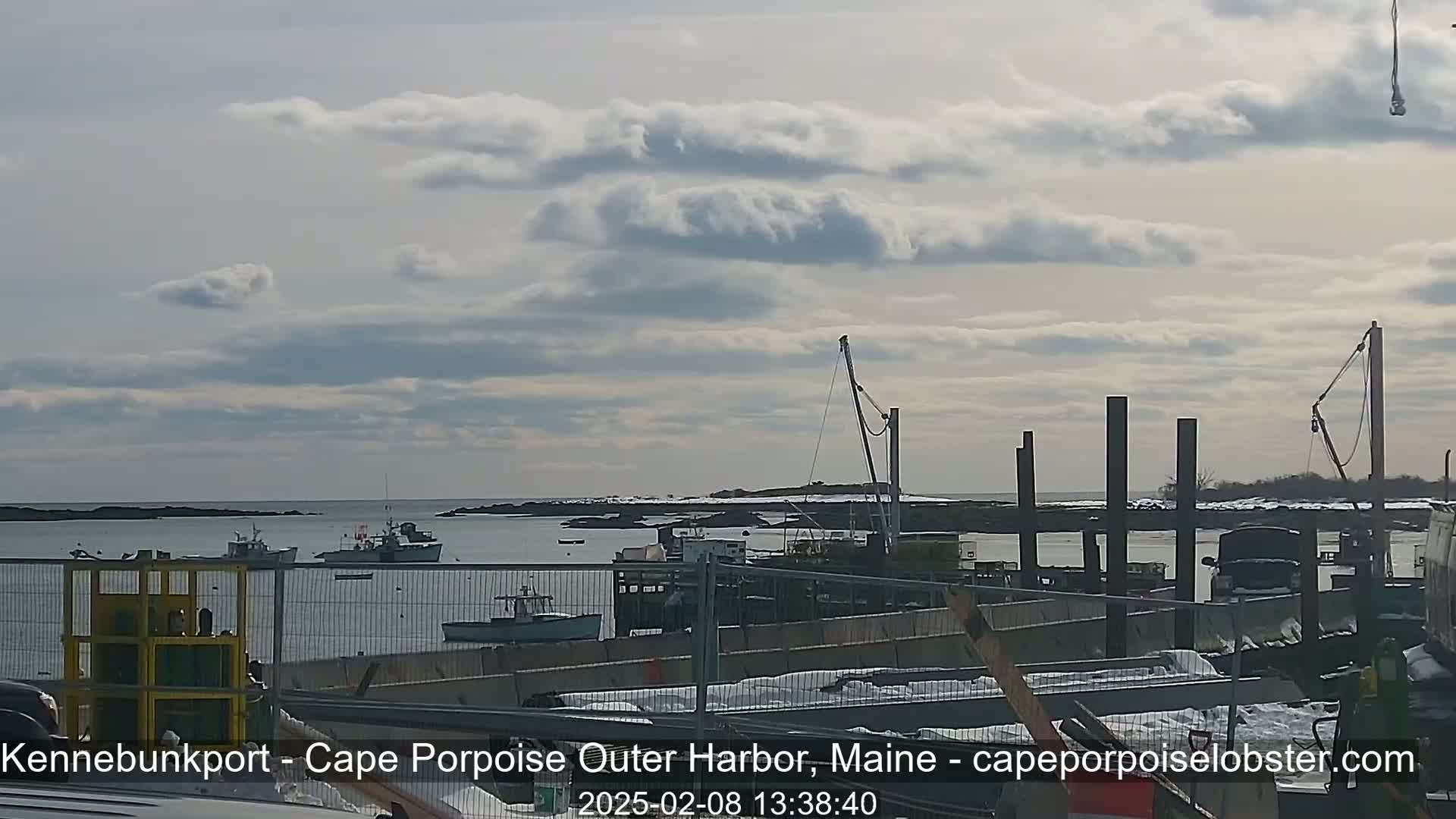 A partly cloudy day reveals a harbor with several boats, construction materials, and a crane, some snow is visible on the land.