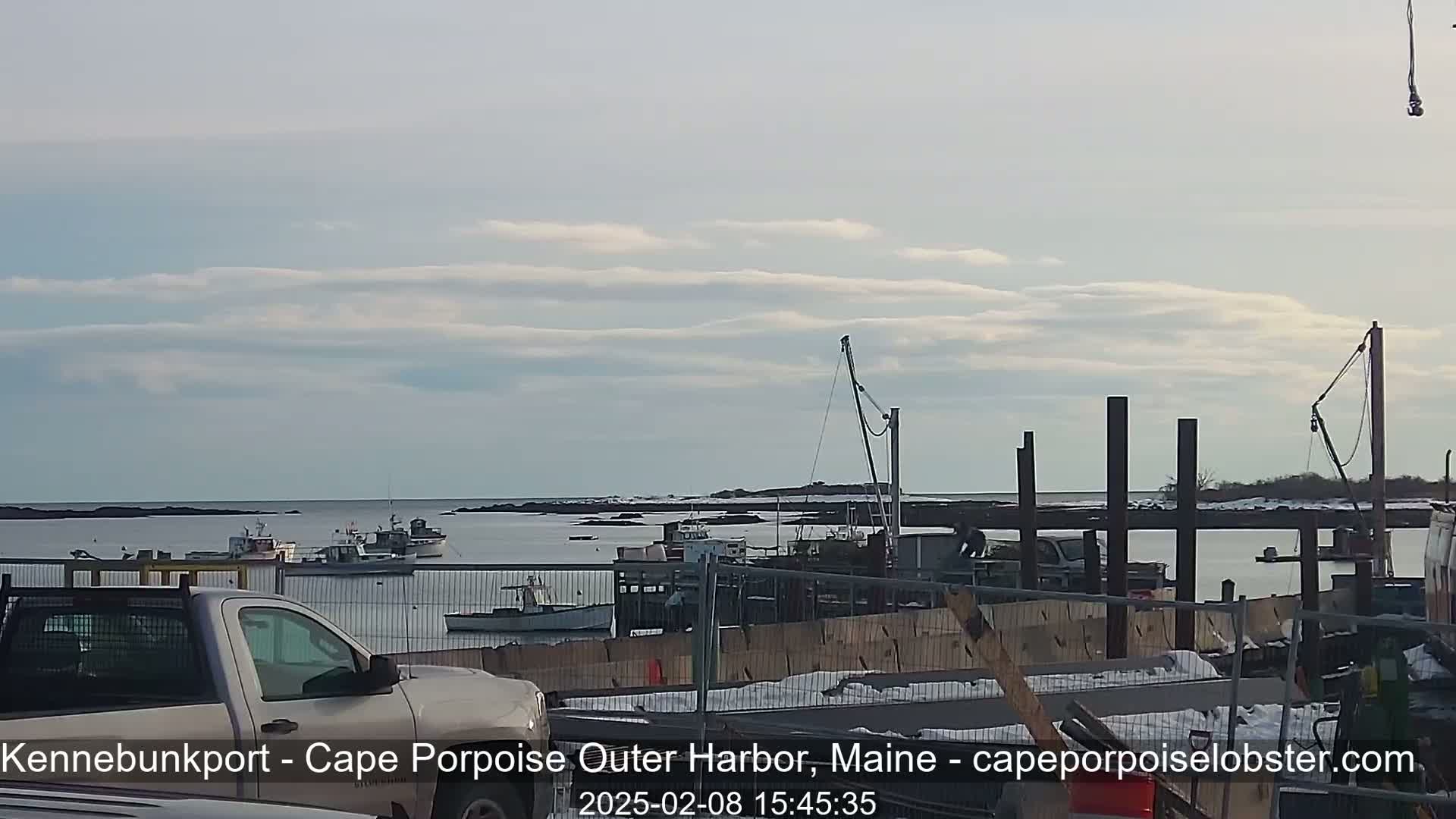 A mostly cloudy day reveals a harbor with several fishing boats, a pickup truck in the foreground, and ongoing construction on a dock.