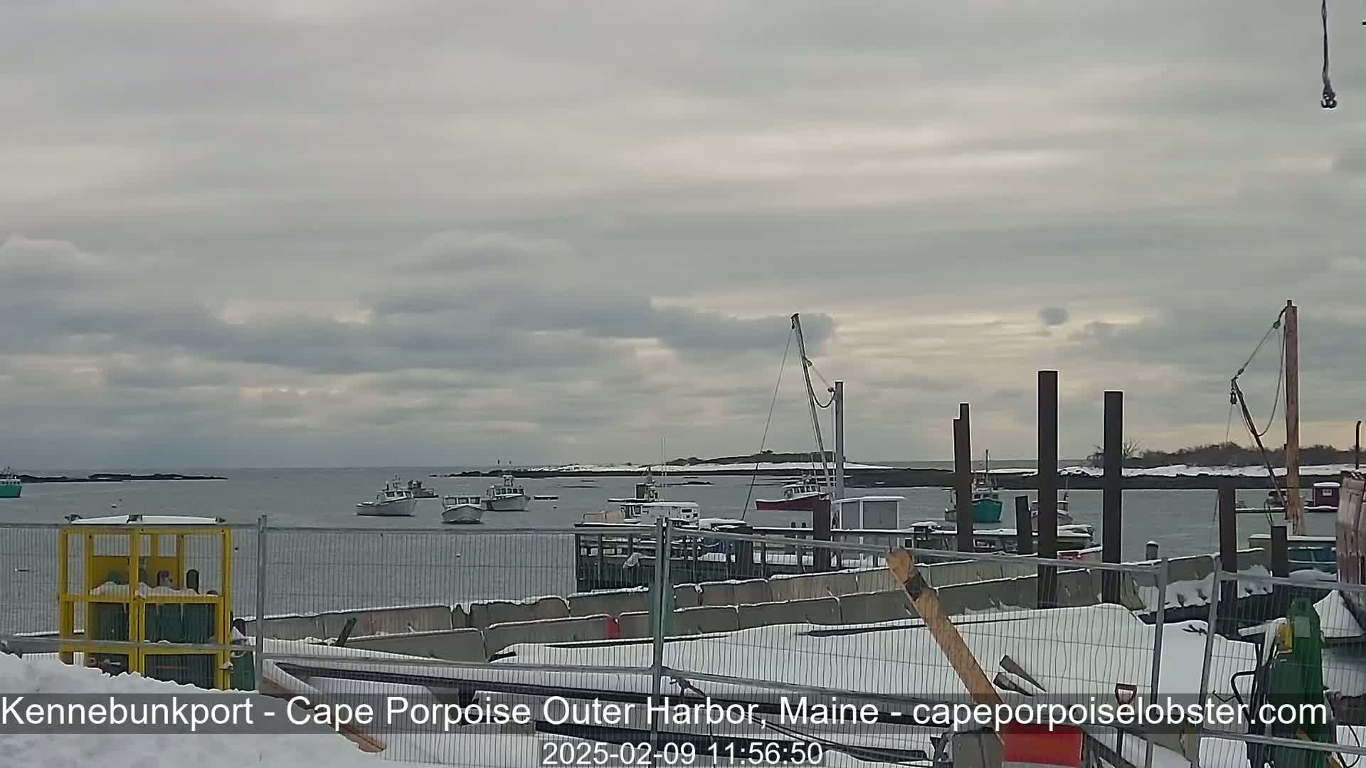 Several boats are moored in a harbor under a cloudy sky, with snow covering the ground and parts of the docks.