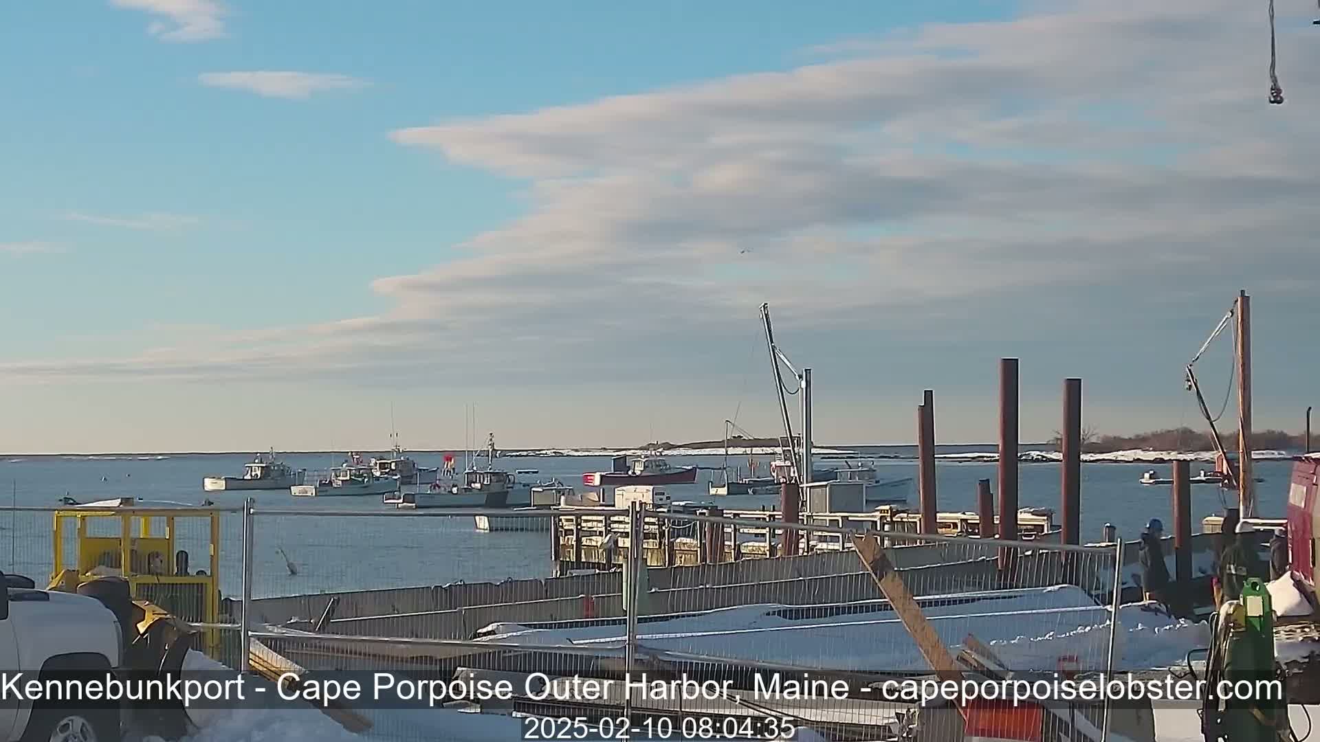 Several fishing boats are moored in a harbor on a partly cloudy day with snow on the ground.