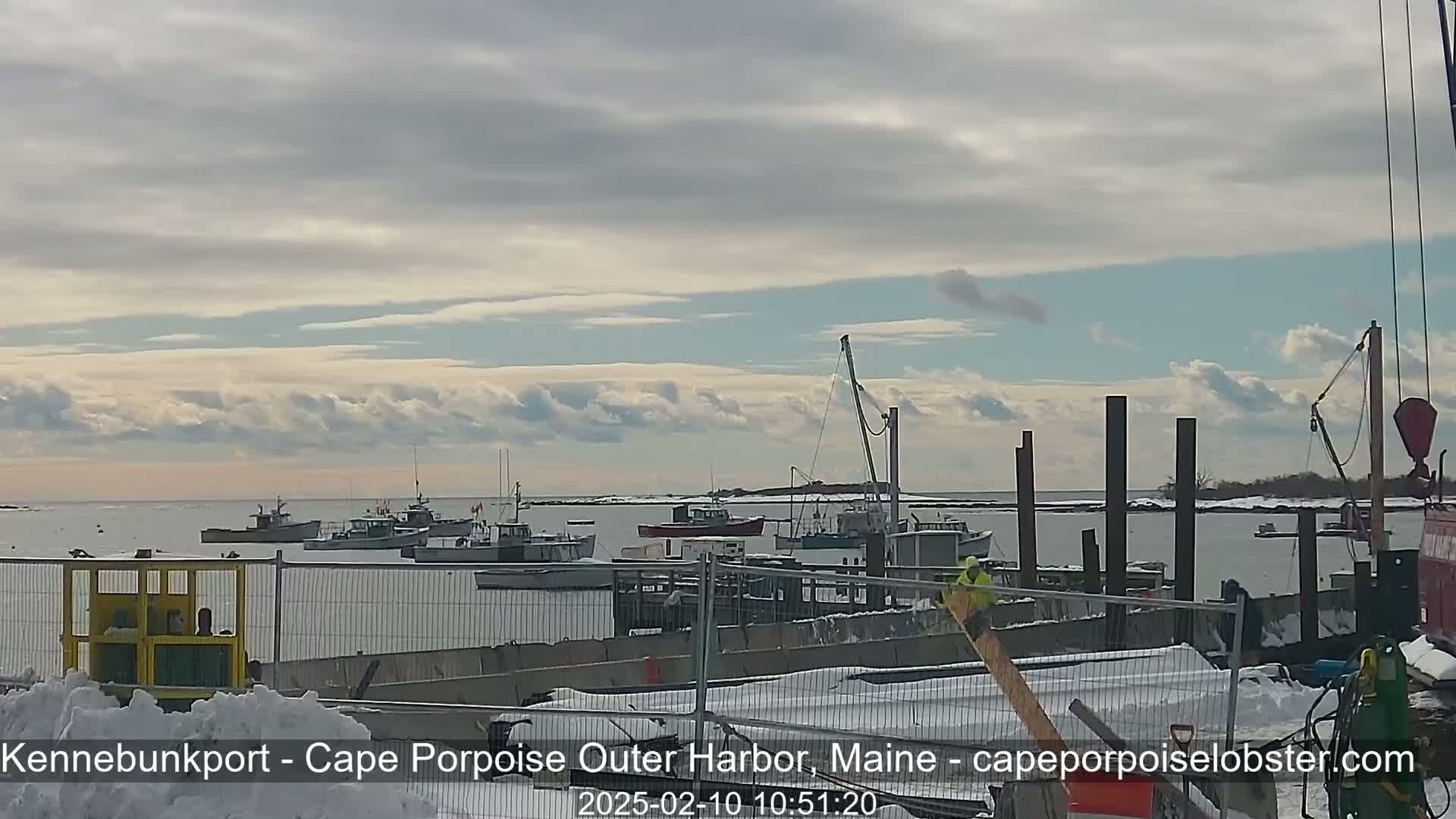 Several fishing boats are moored in a harbor under a mostly cloudy sky, with snow on the ground and a construction site visible in the foreground.