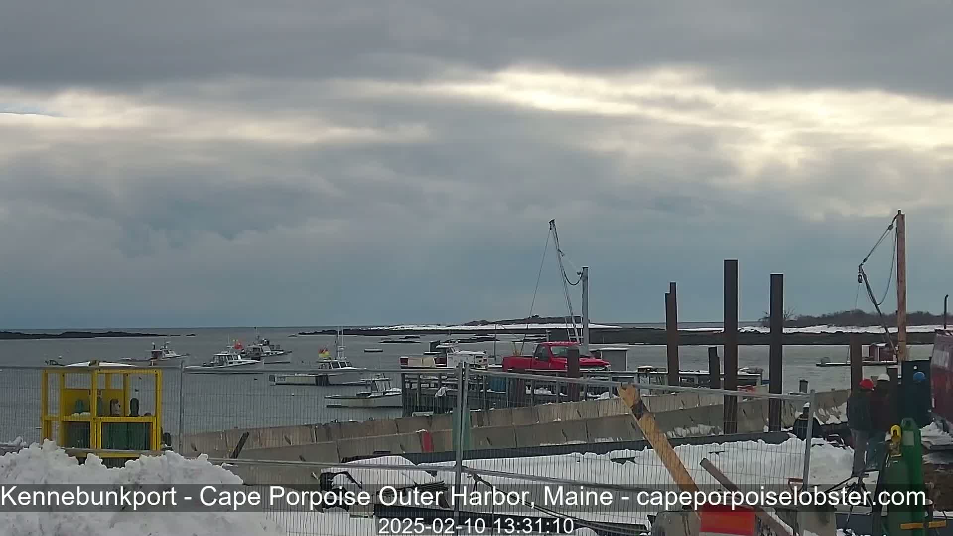 Several boats are moored in a harbor under a cloudy sky, with snow on the ground and ongoing construction visible in the foreground.