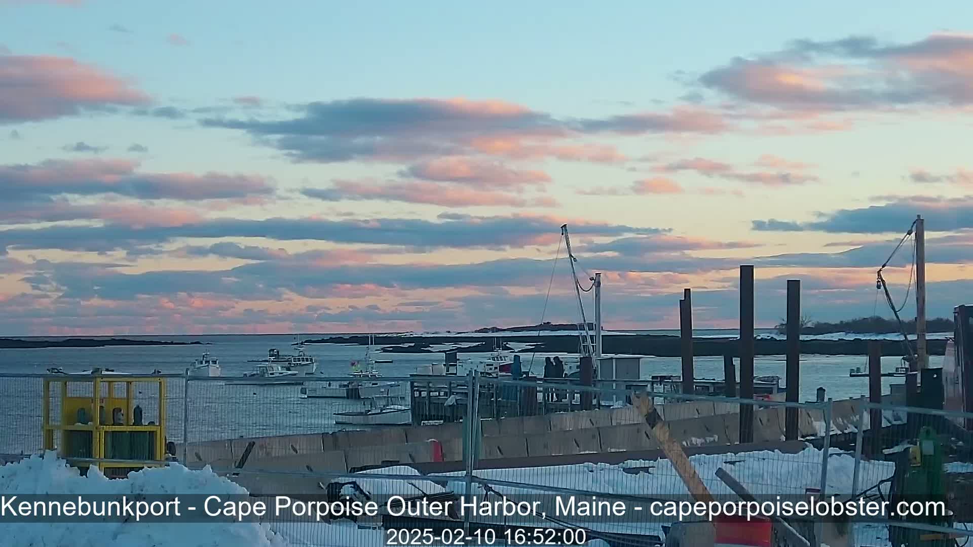 Several boats are moored in a harbor under a partly cloudy sky with pink and blue hues at sunset, and there is snow on the ground near a construction site.