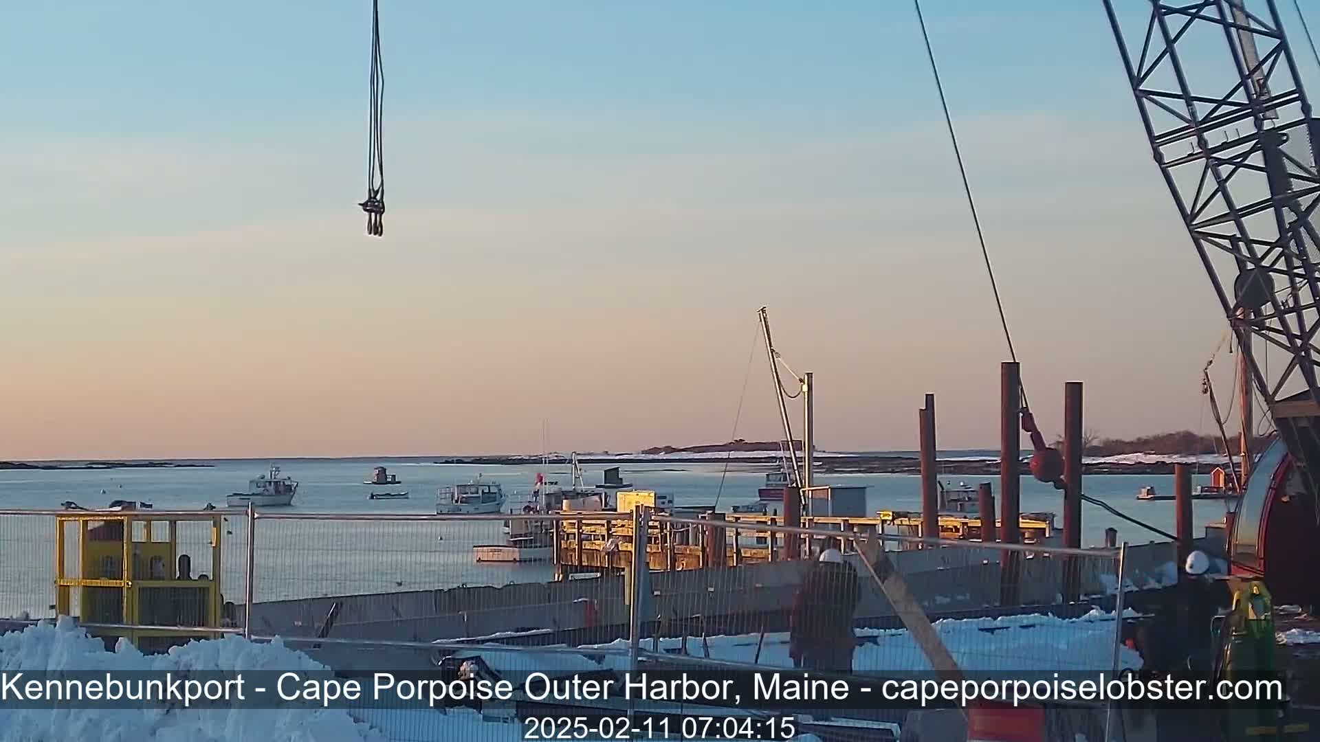 Several boats are moored in a harbor under a pale pink sunset sky, with a crane and construction in the foreground and some snow on the ground.