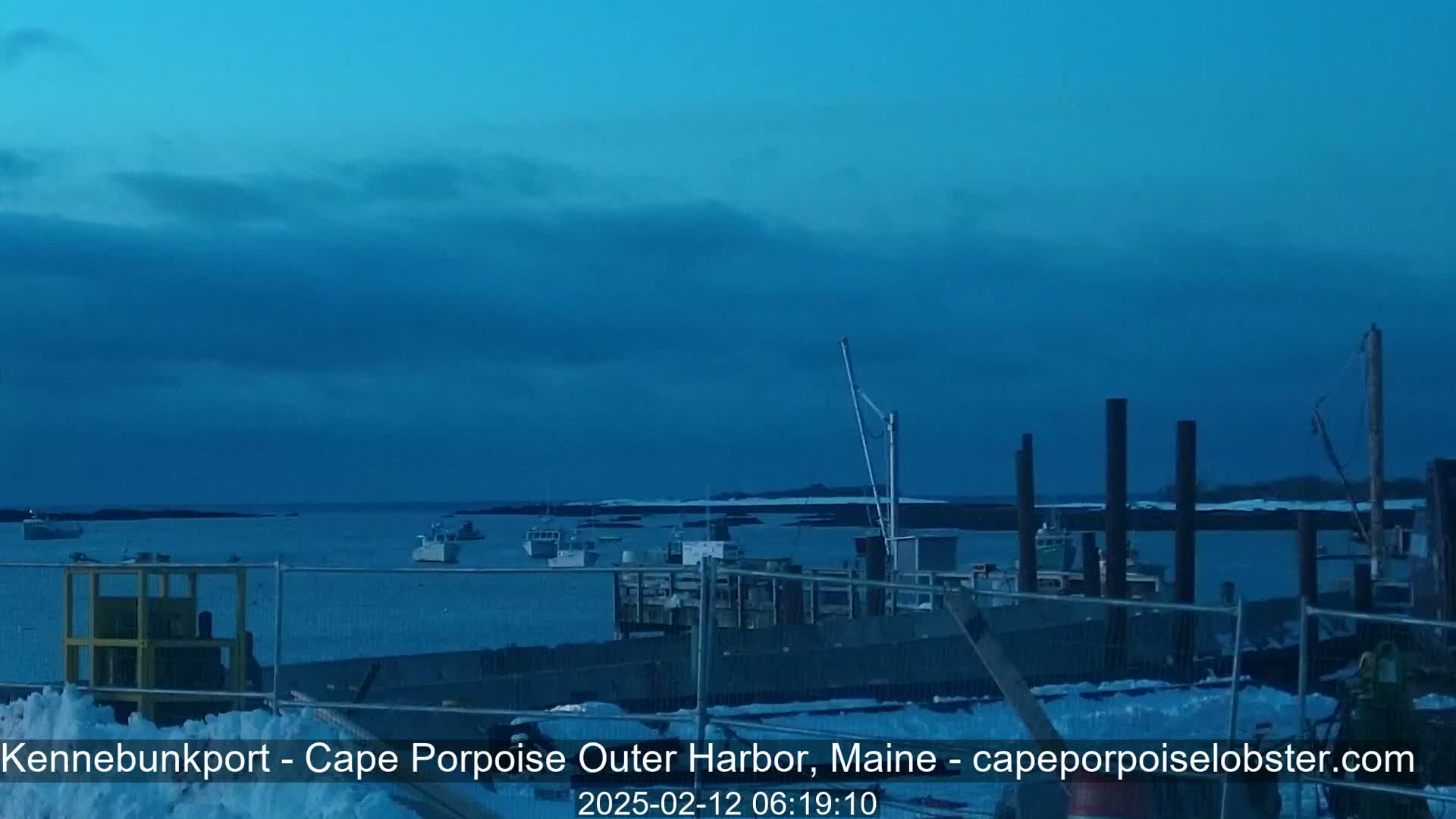 A mostly cloudy, dark blue twilight sky overlooks a harbor with several boats moored at a dock partially covered in snow.