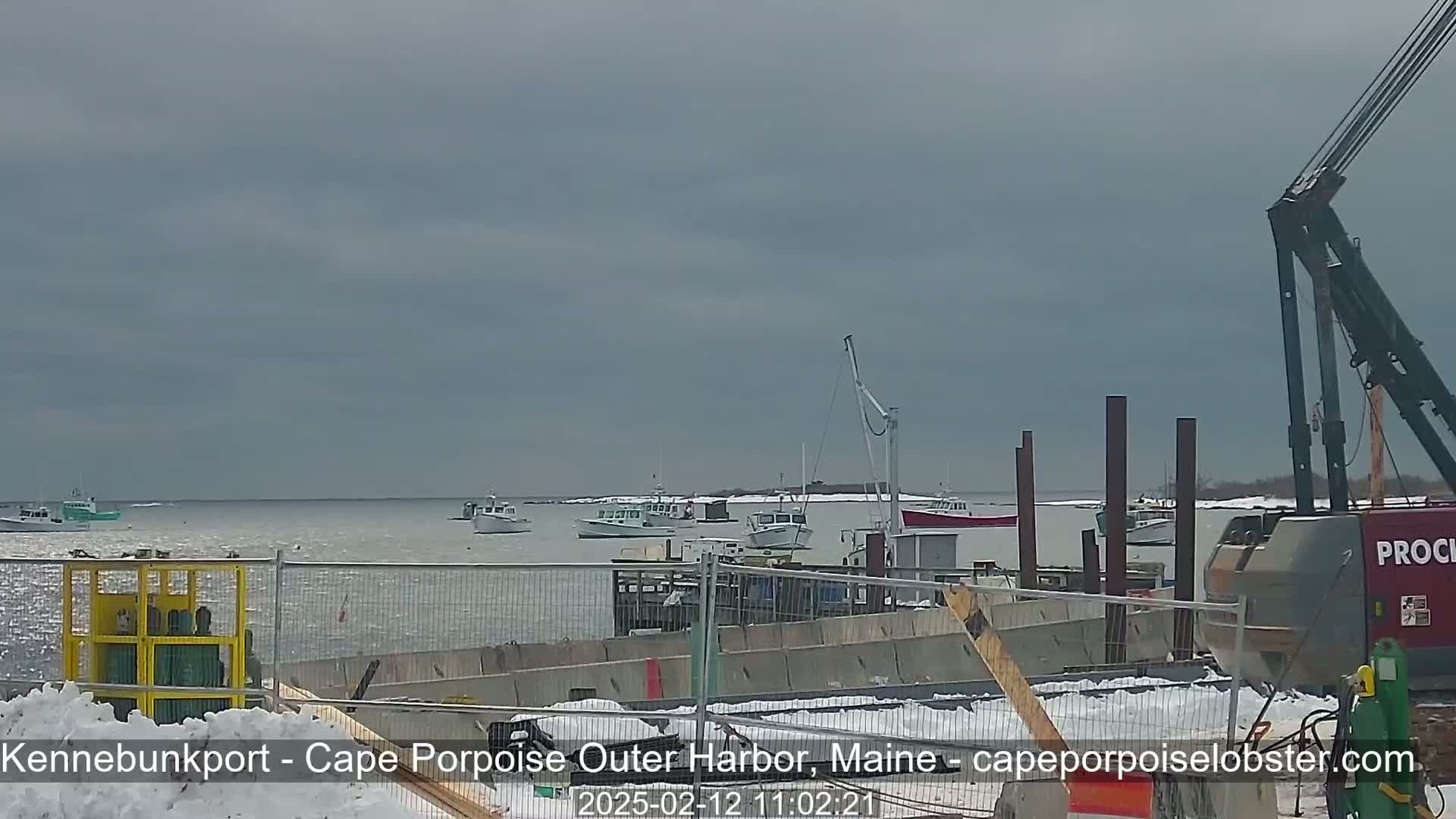 Several fishing boats are moored in a harbor on an overcast day with patches of snow on the ground and a construction crane in the foreground.