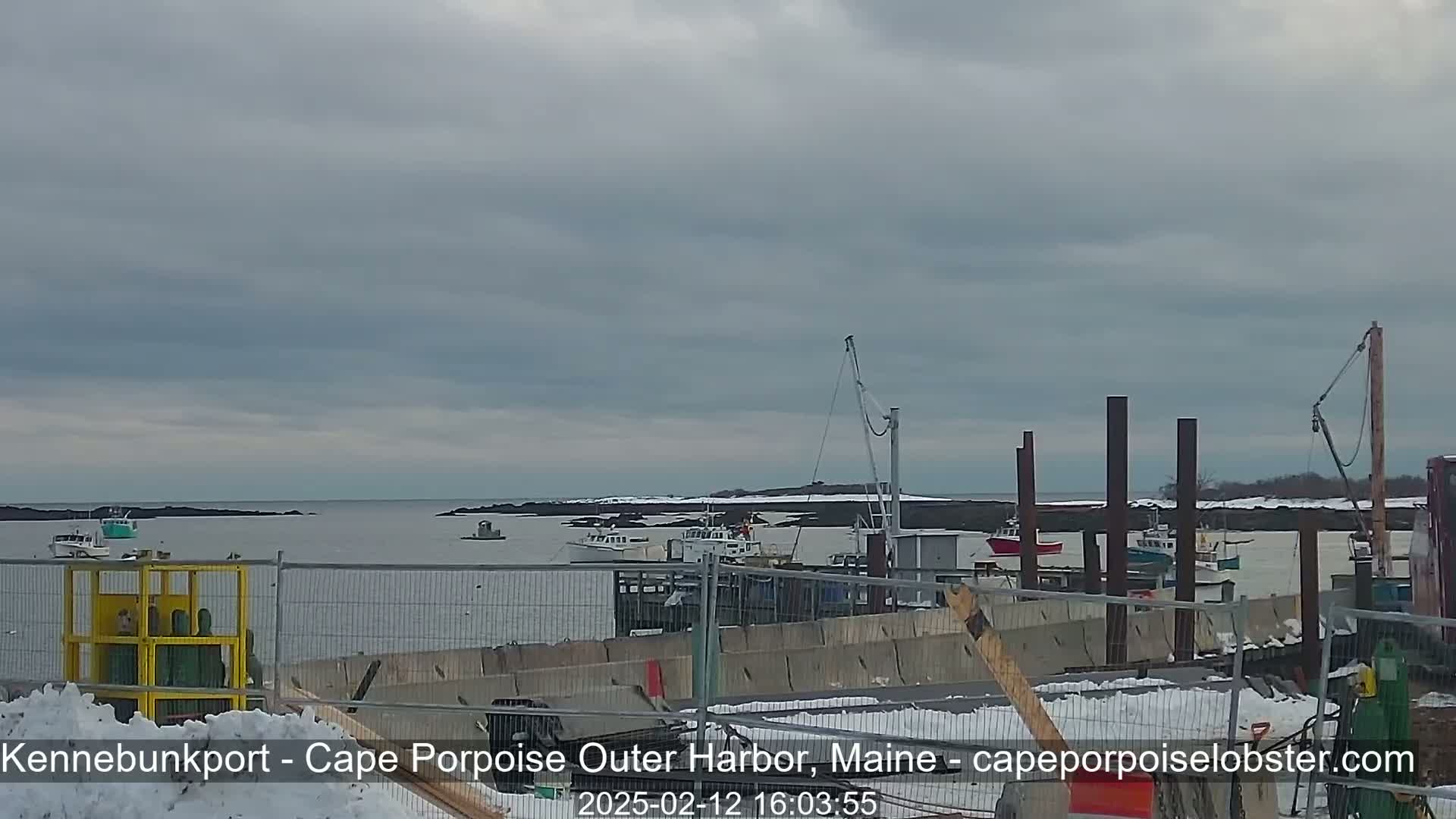 Several fishing boats are moored in a harbor under a cloudy sky, with some snow on the ground near a construction site.