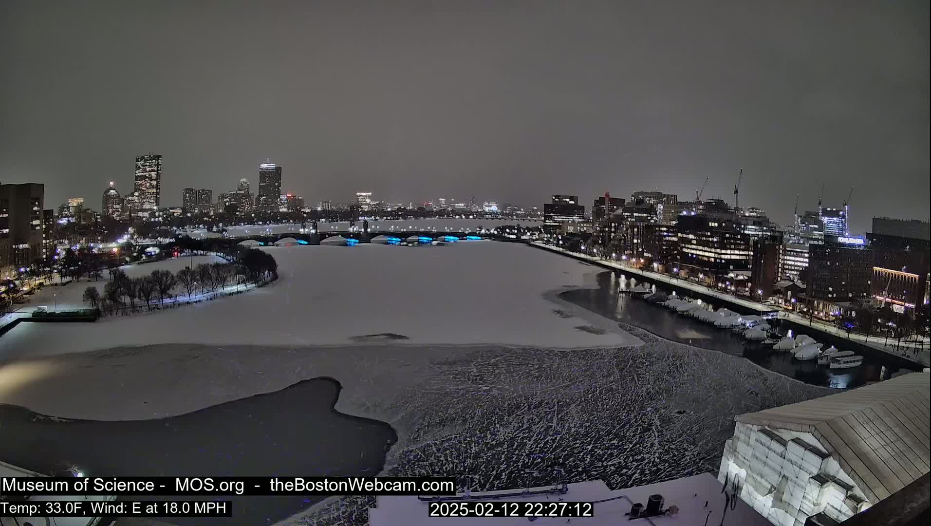 A nighttime cityscape with a partially frozen river, snow-covered ground, and an 18 mph east wind at 33°F.