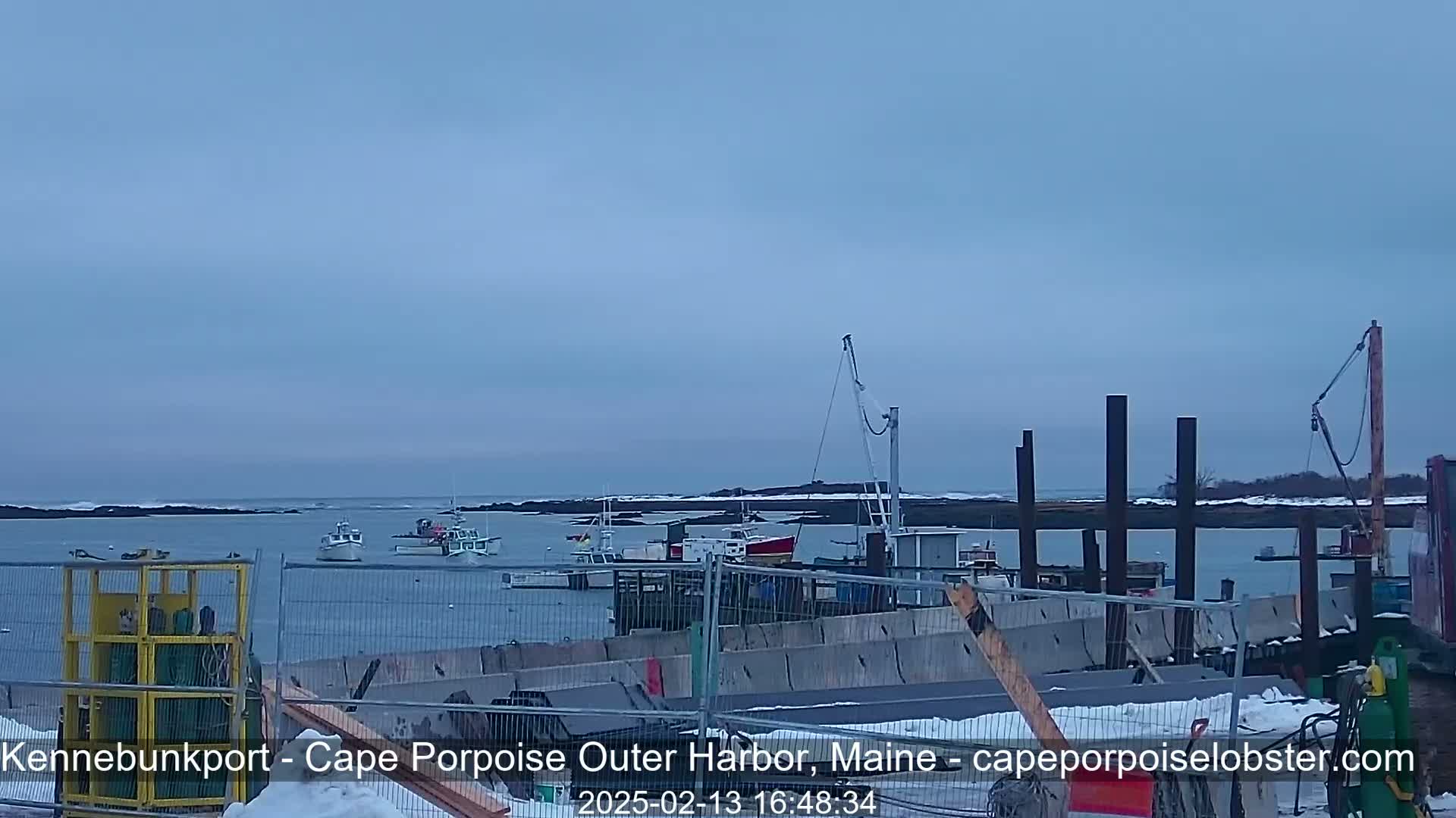 Several small fishing boats are moored in a harbor under an overcast sky, with some construction or repair work visible in the foreground.