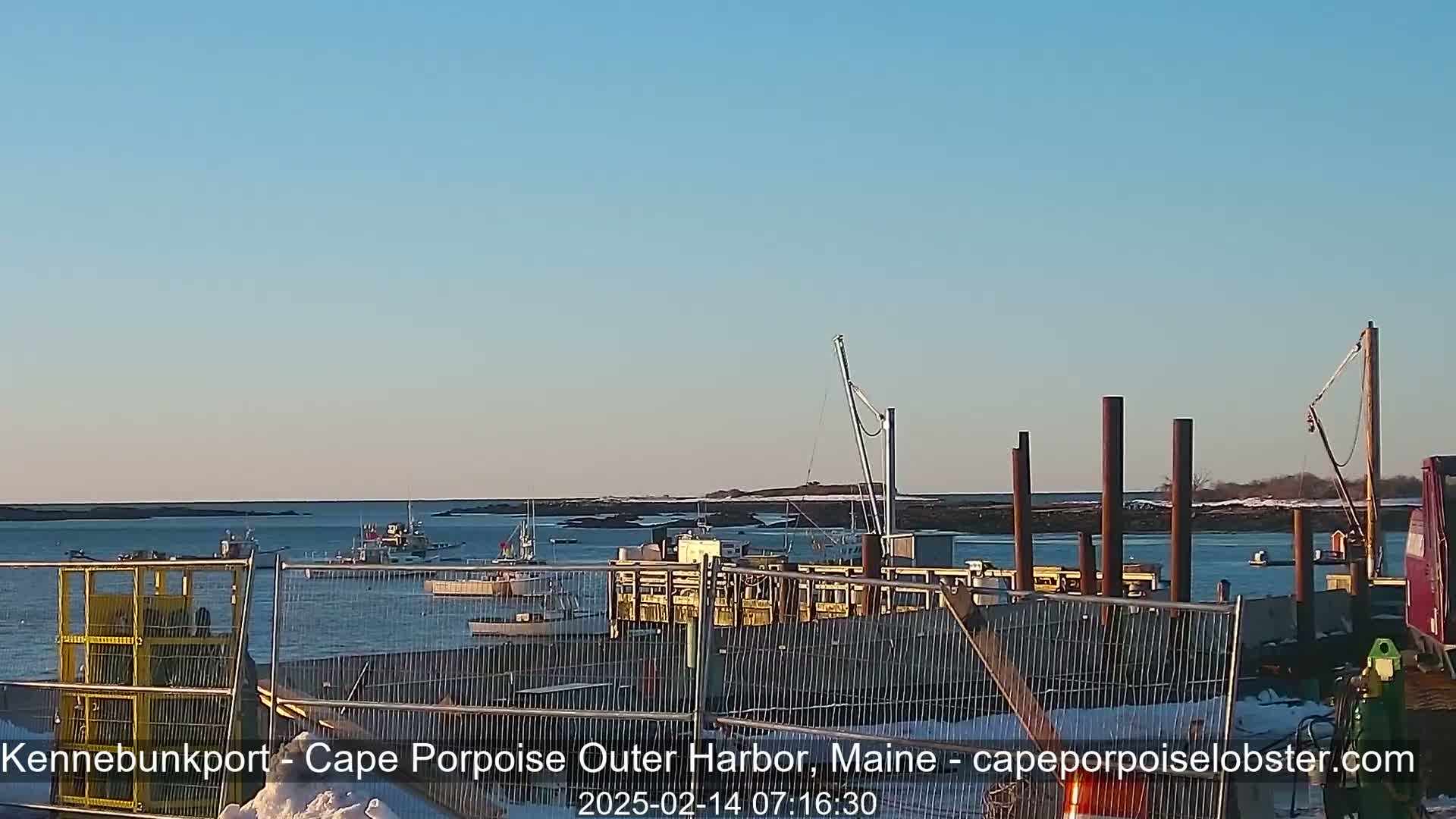 A harbor with several fishing boats is partially obscured by a metal fence and construction materials under a clear, bright sky.