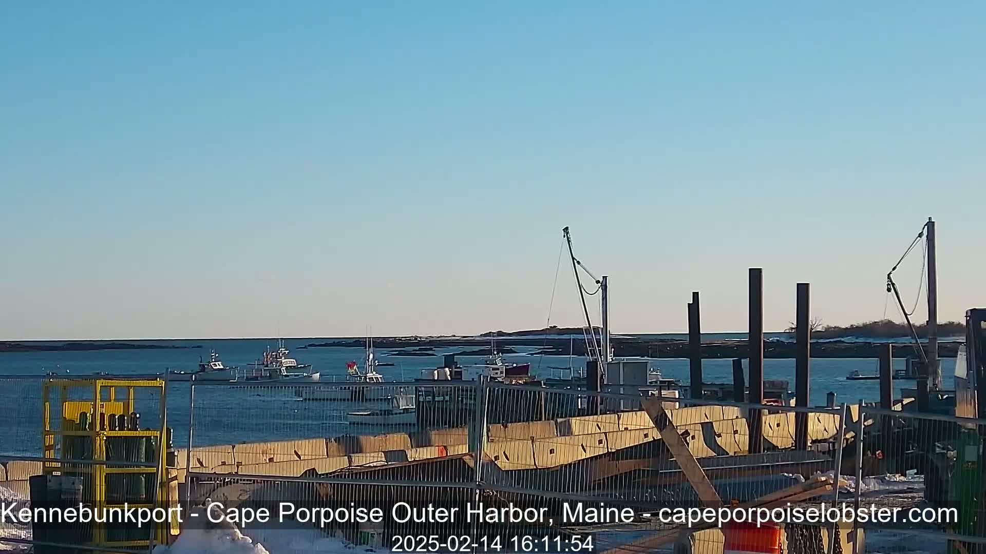 Several fishing boats are moored in a harbor under a clear blue sky, with construction materials and fencing visible in the foreground.