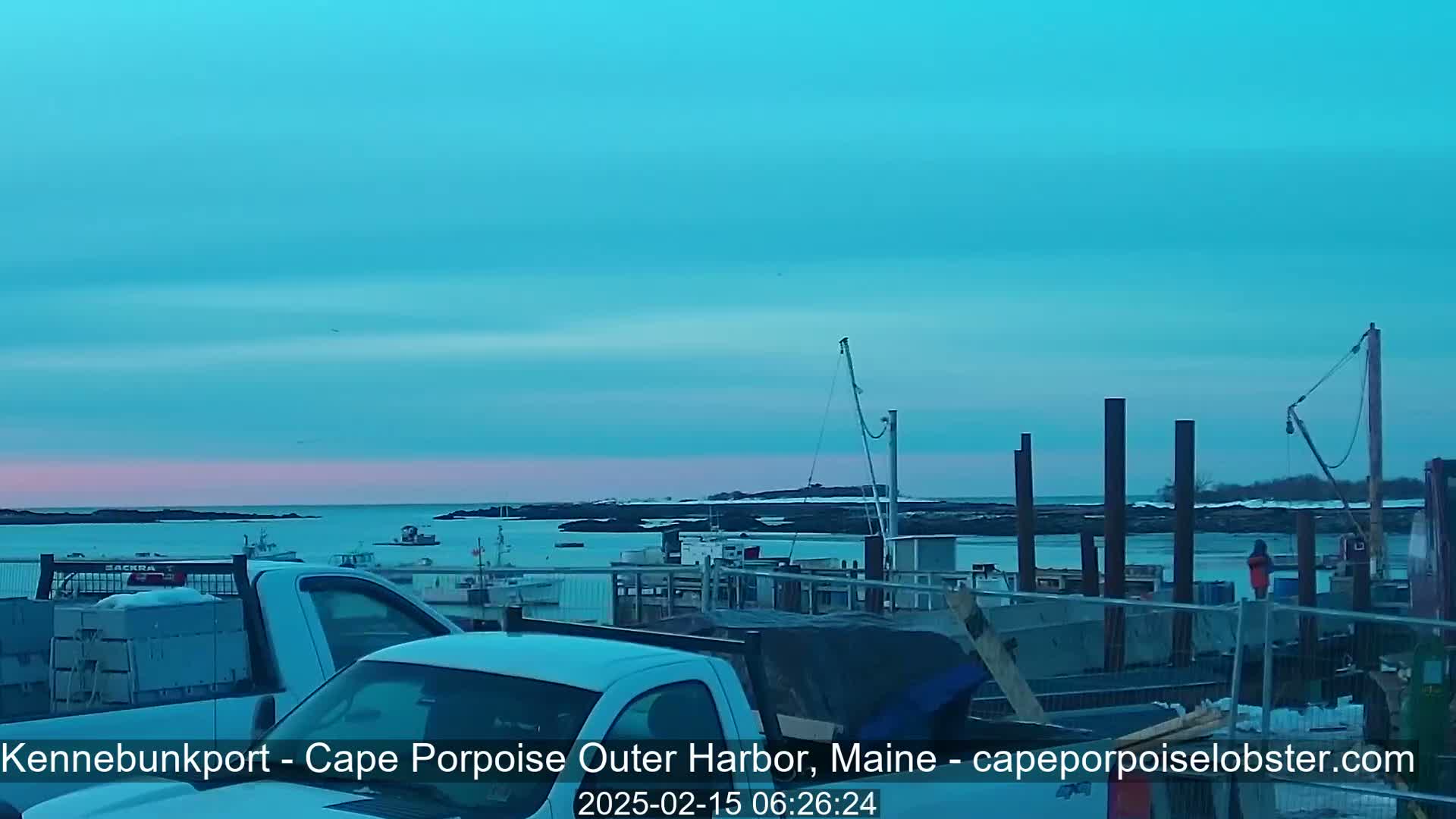 A tranquil harbor scene at dawn shows several small fishing boats moored near a rocky shoreline under a pale blue sky.