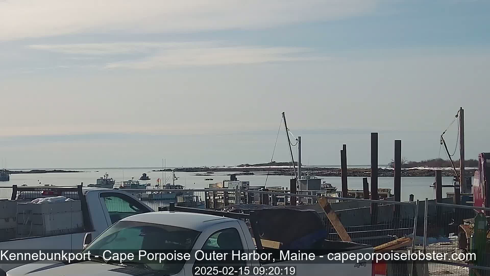 Several boats are docked in a harbor on a partly sunny day, with some trucks and construction materials visible in the foreground.