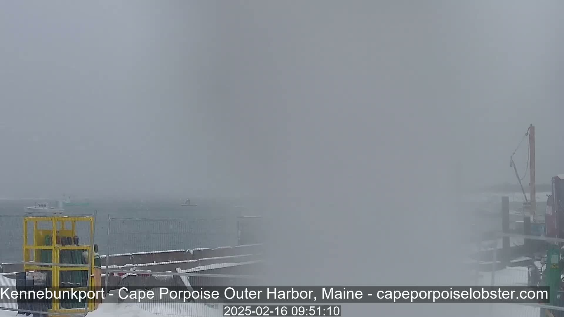 A snow-covered harbor scene is visible through heavy fog or blowing snow, with a few boats on the water and some construction equipment on the shore.