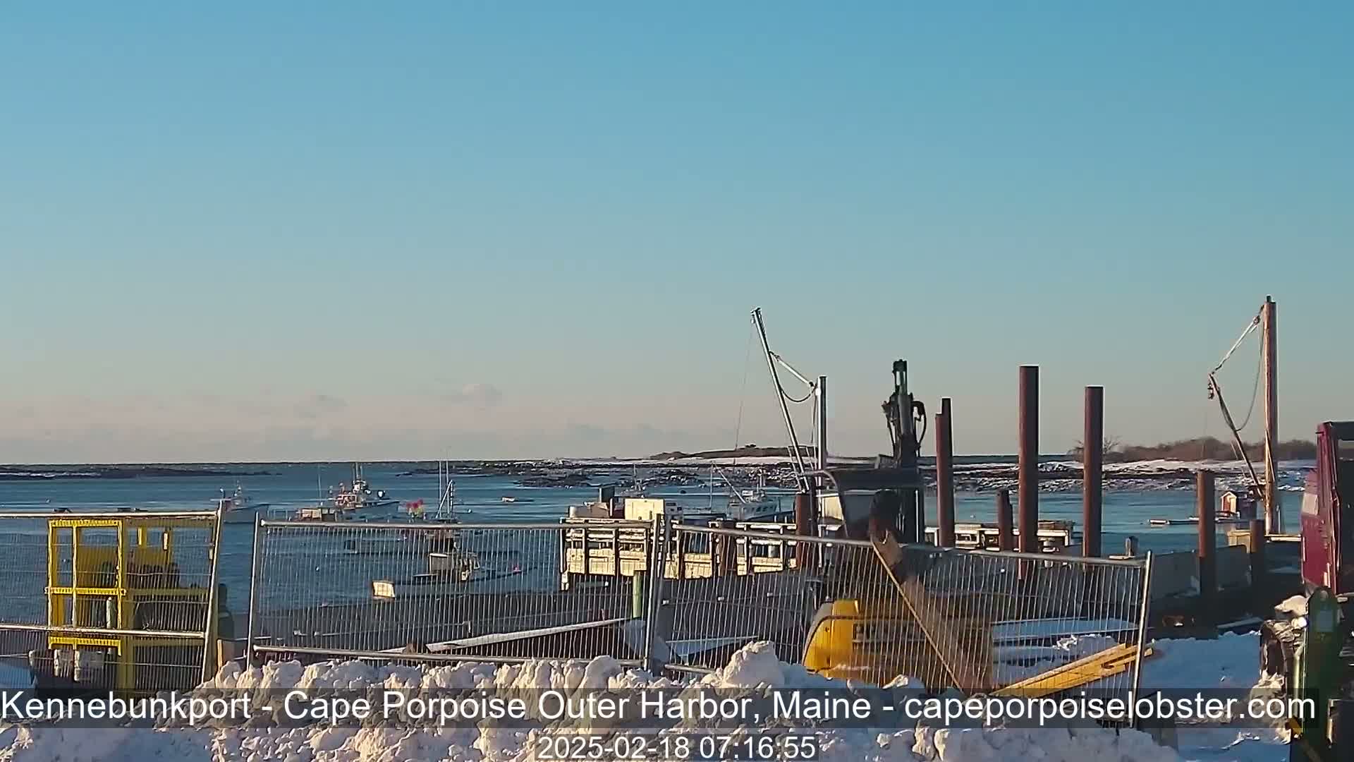 A clear, sunny day shows a harbor with several fishing boats, a dock under construction, and snow on the ground.