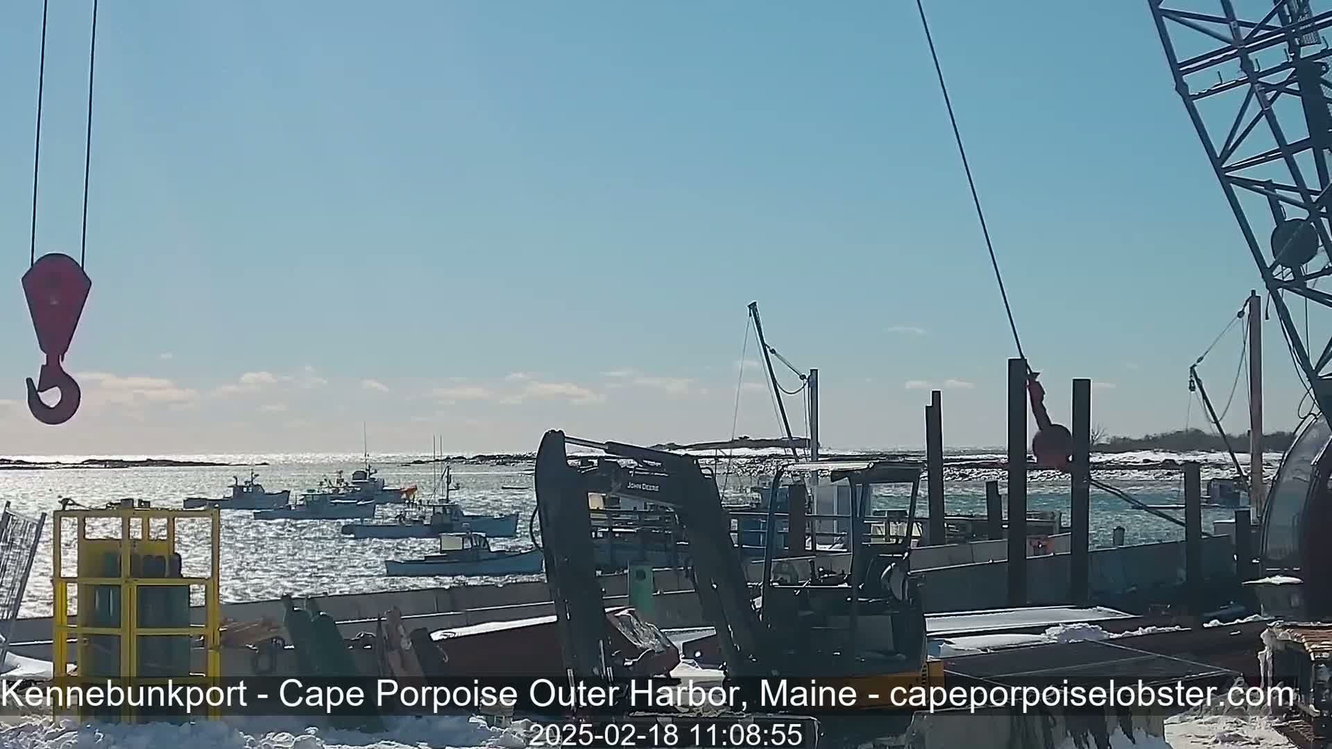 A harbor with several fishing boats is viewed from a dock on a sunny, cold day with some snow visible on the ground and ice on the water.