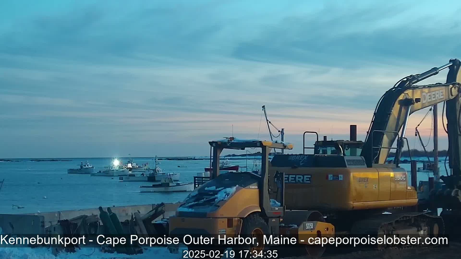 Several fishing boats are moored in a harbor at twilight under a mostly clear sky, with heavy construction equipment parked nearby.