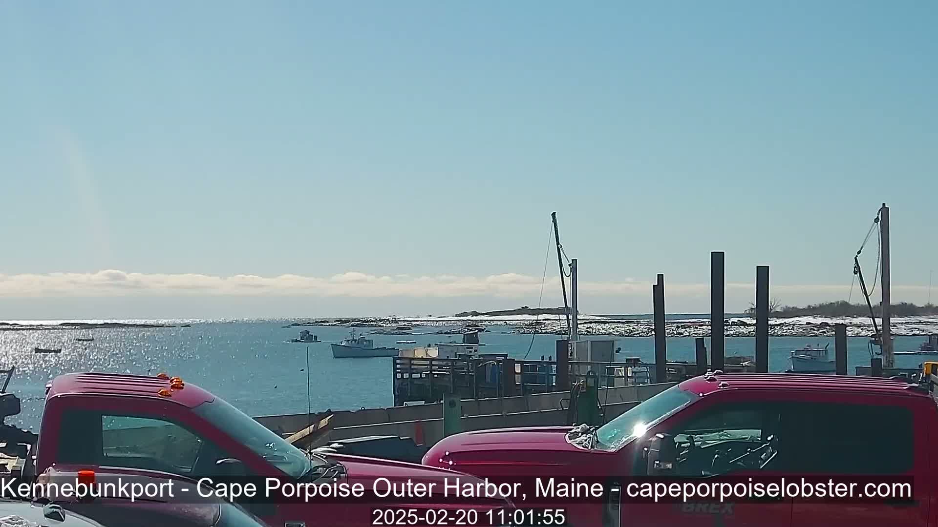 Two red pickup trucks are parked in front of a harbor with several boats and a partially snowy shoreline under a mostly sunny sky.