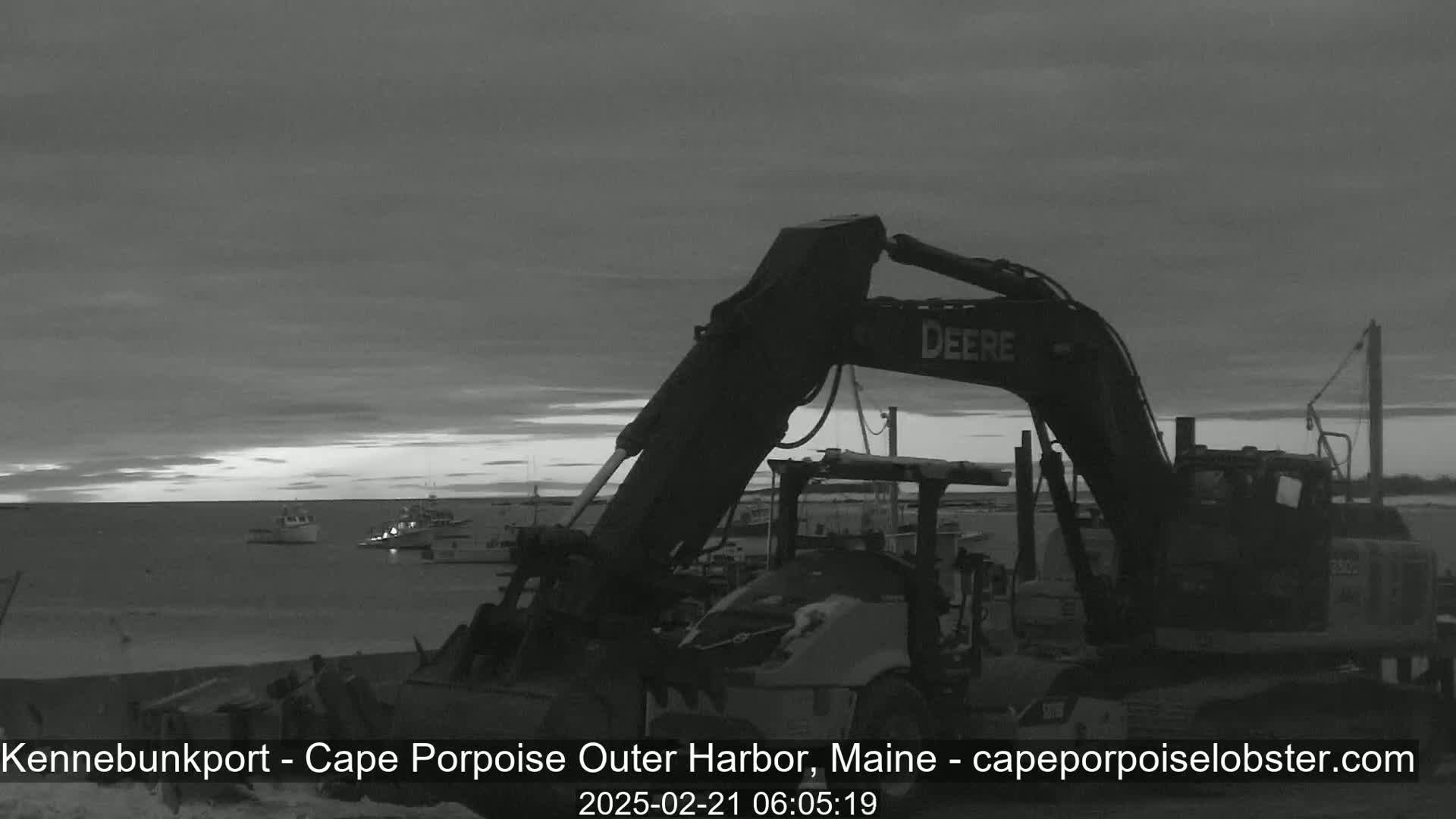 A large excavator sits on a harbor shore at dawn under an overcast sky, with several boats visible on the water.