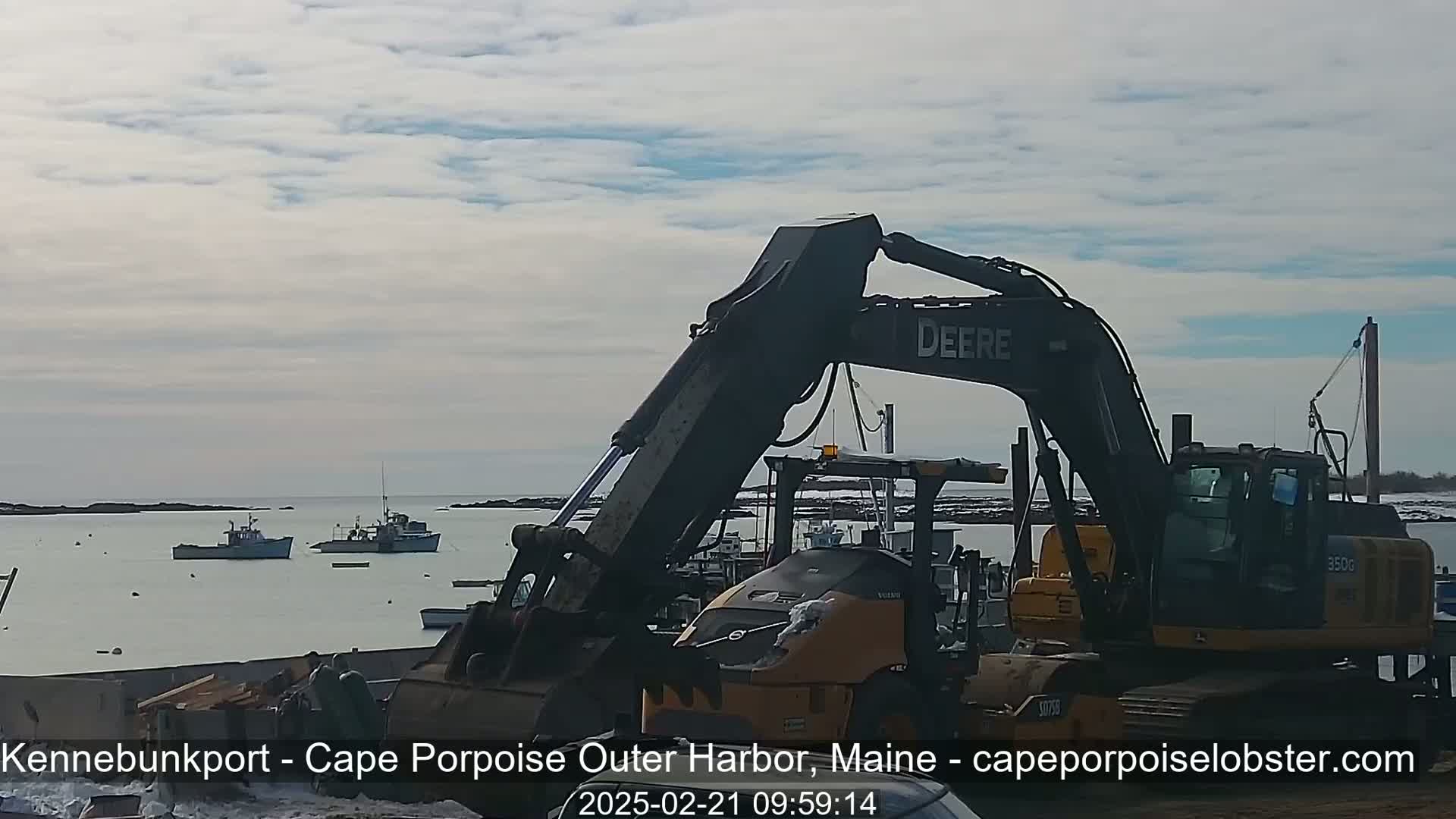 A large excavator sits in a harbor next to several fishing boats on a partly cloudy day.