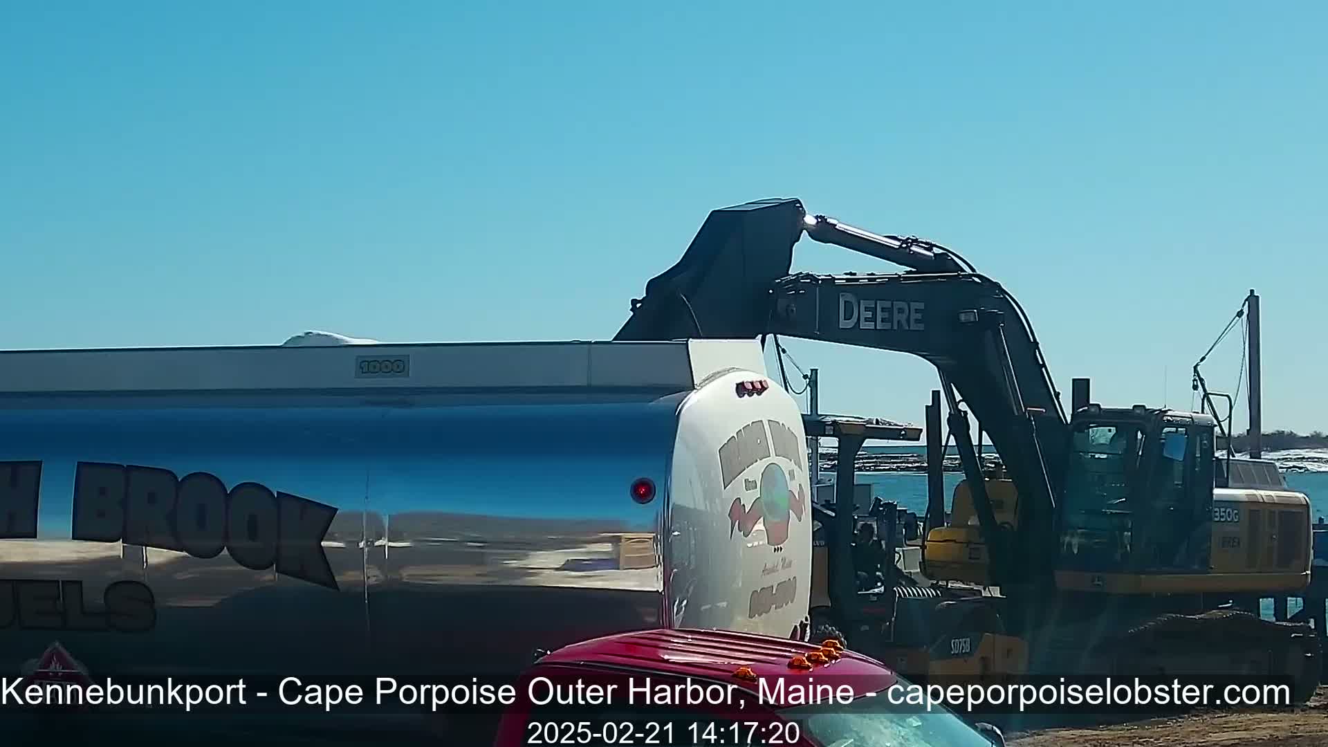 A silver fuel tanker truck is being filled by a Deere excavator on a sunny day near a body of water.