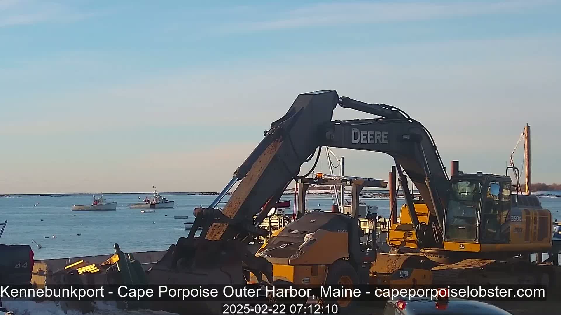 A large excavator and a smaller road roller are positioned near a harbor with several fishing boats in the water under a clear, sunny sky.